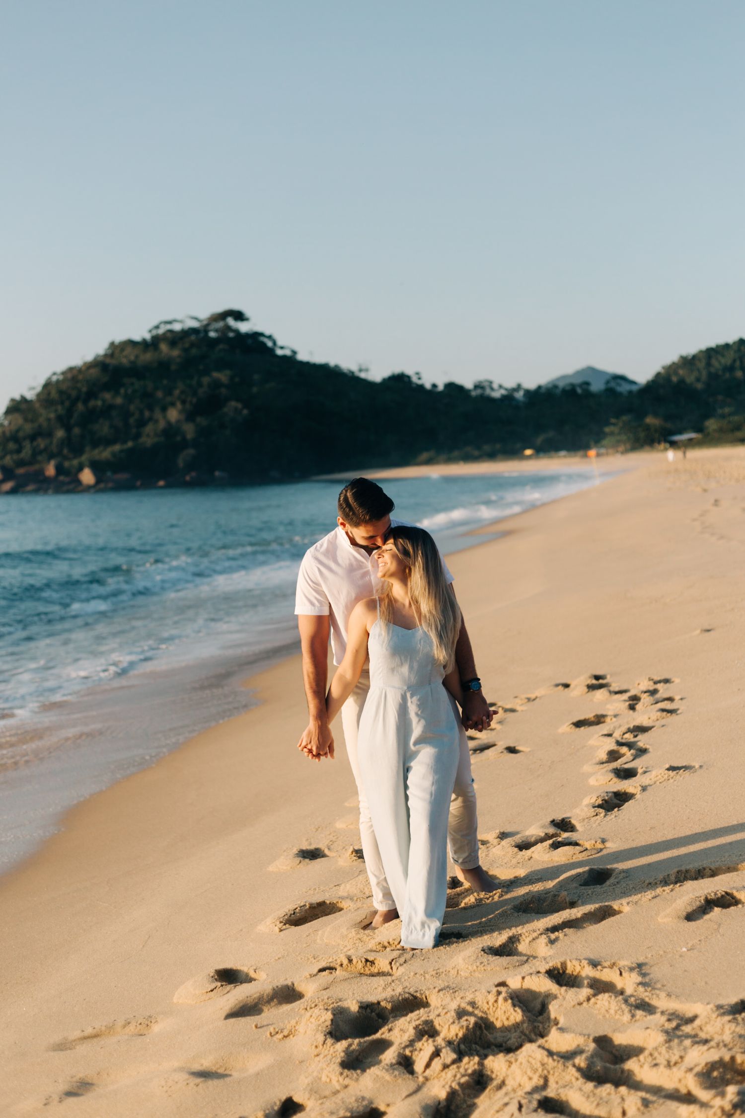 Couple dressed in white sharing a tender moment while walking along a scenic coastal beach.