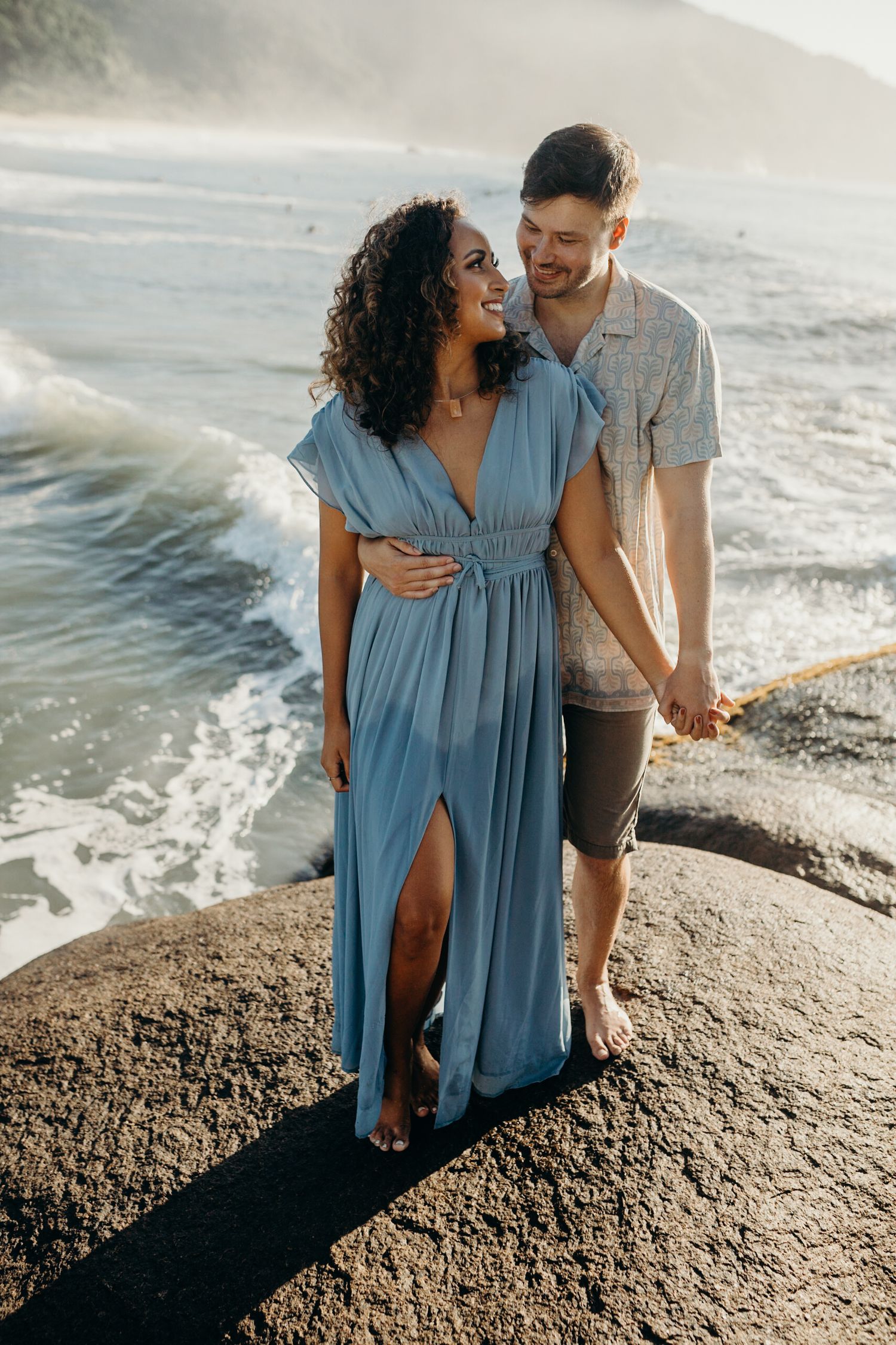Couple poses together on large rock overlooking scenic beach during golden hour.