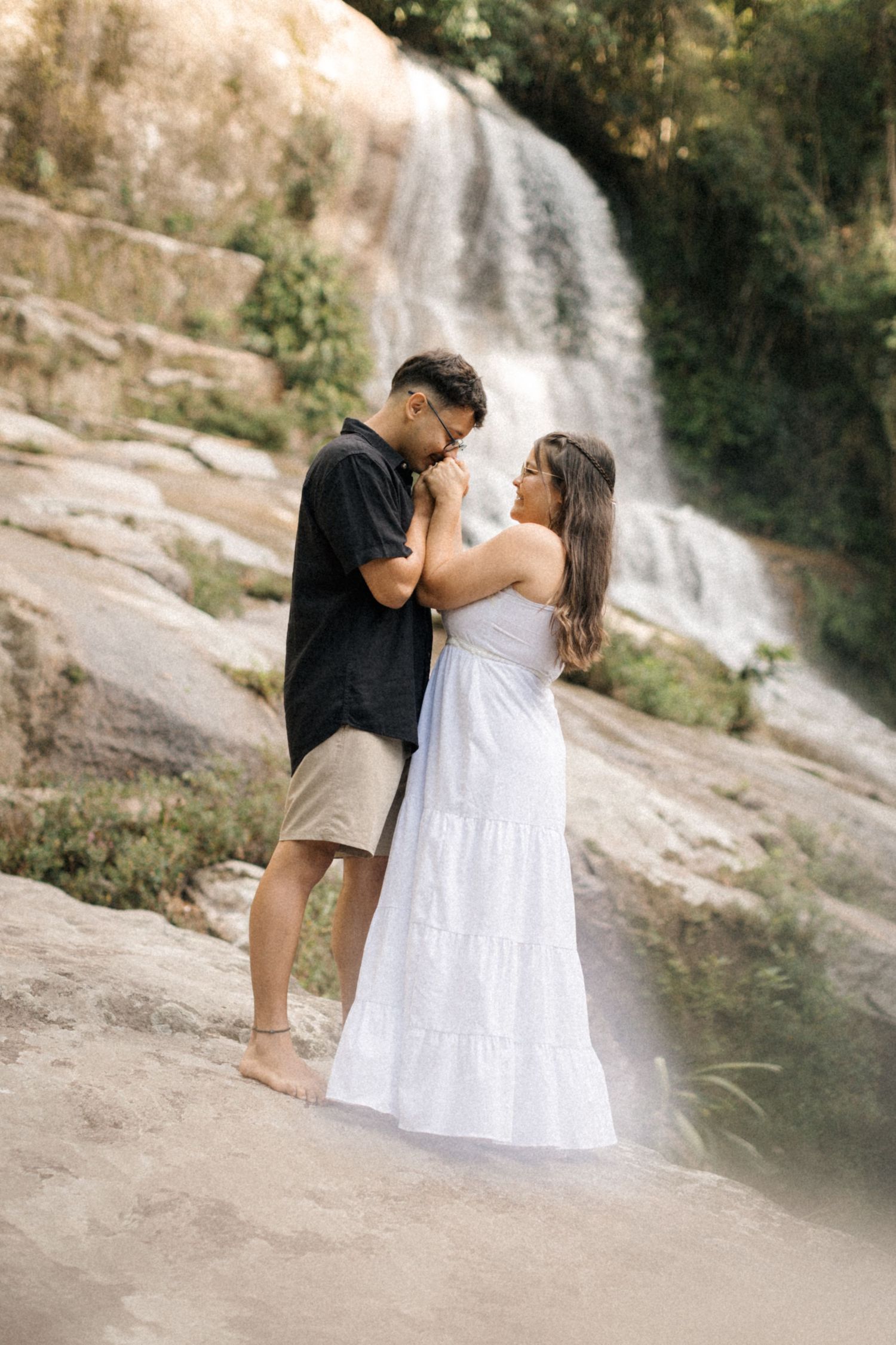 A romantic photoshoot featuring a couple in casual attire against a waterfall backdrop.