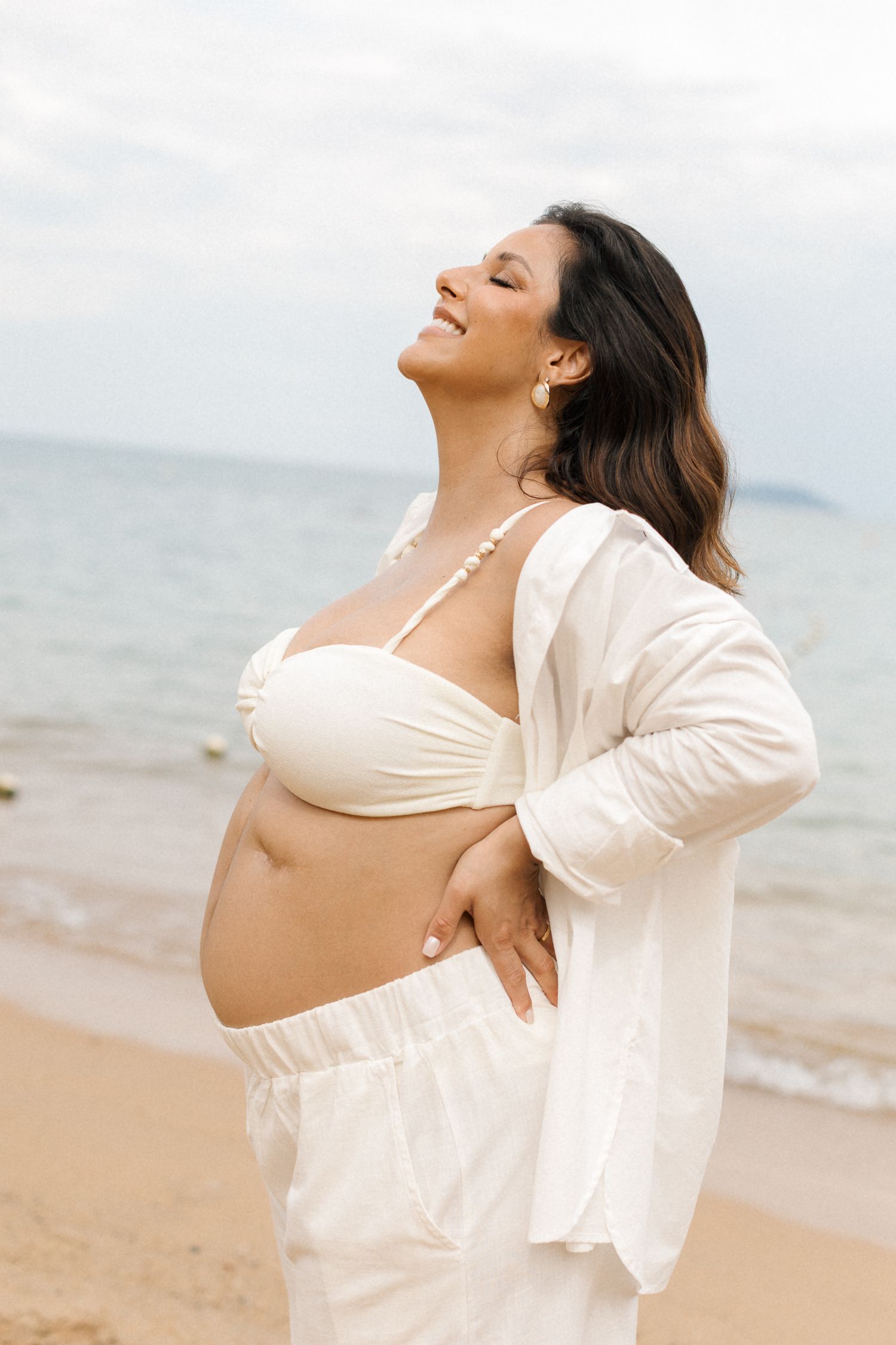 Expectant mother in white beachwear stands joyfully on sandy beach with ocean waves in background.