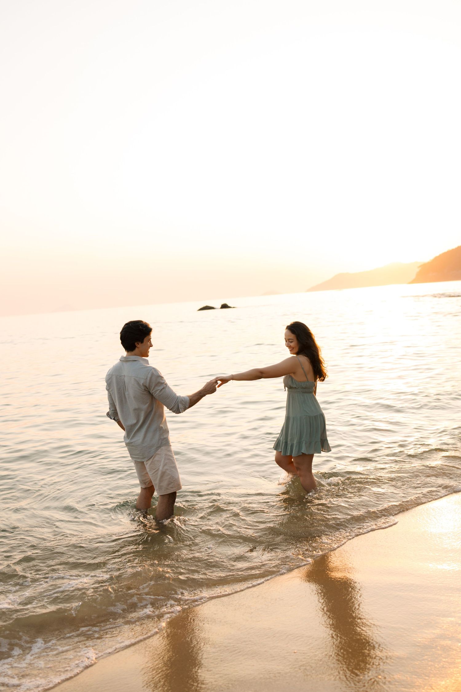 Couple holds hands while wading in shallow ocean water during golden hour sunset at beach.