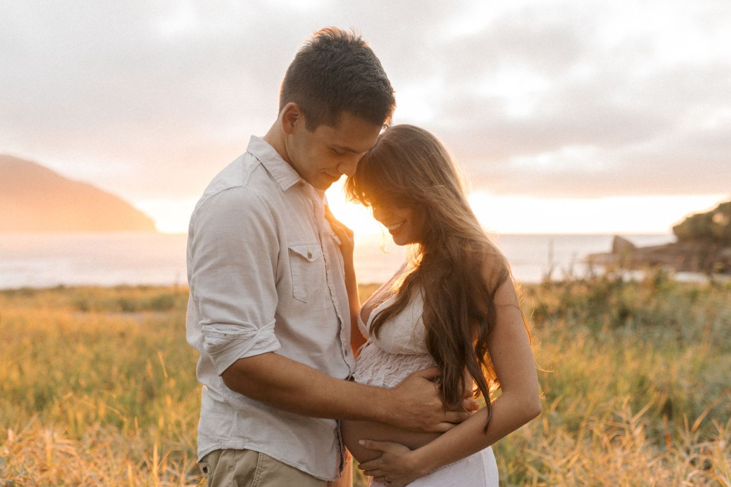 A couple embraces in a golden field during sunset while wearing white and beige outfits for their engagement photos.
