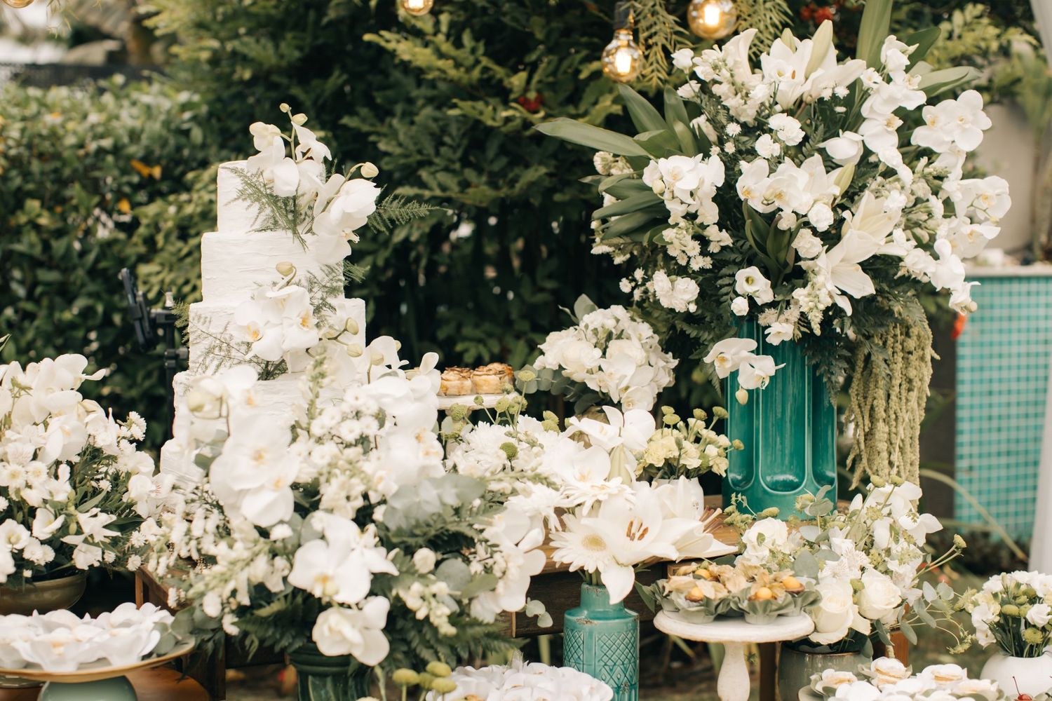 White floral wedding cake display surrounded by lush flower arrangements and greenery.