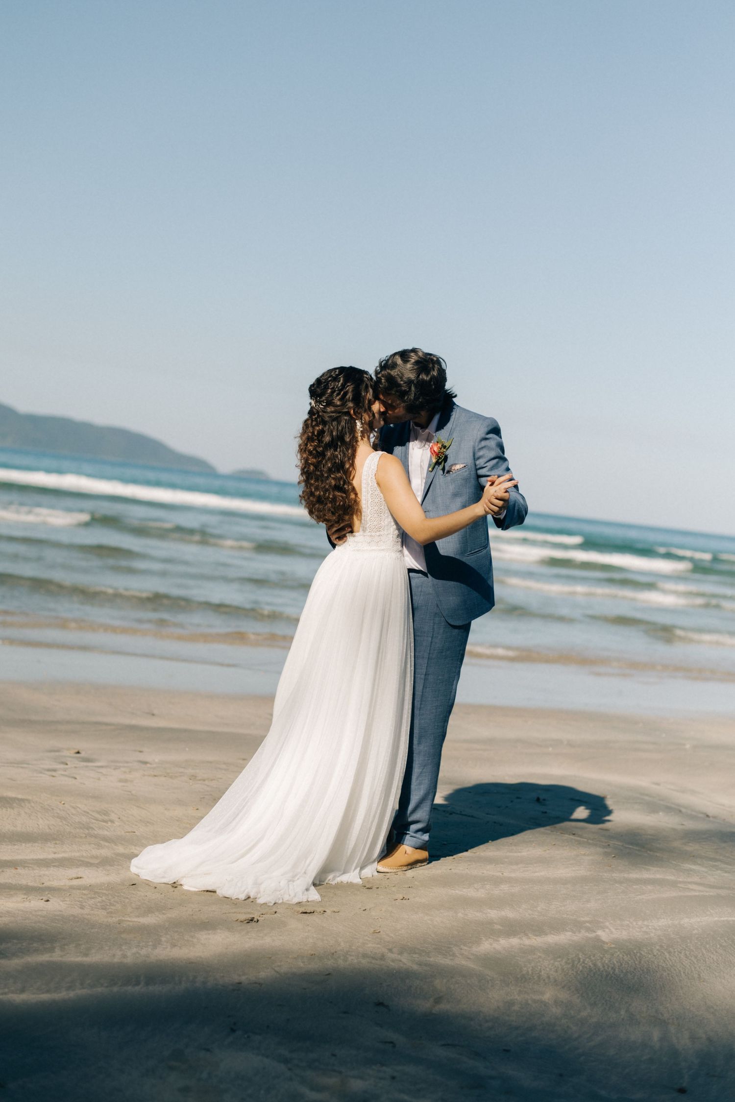 Intimate beach wedding moment captured with ocean waves and clear blue sky.
