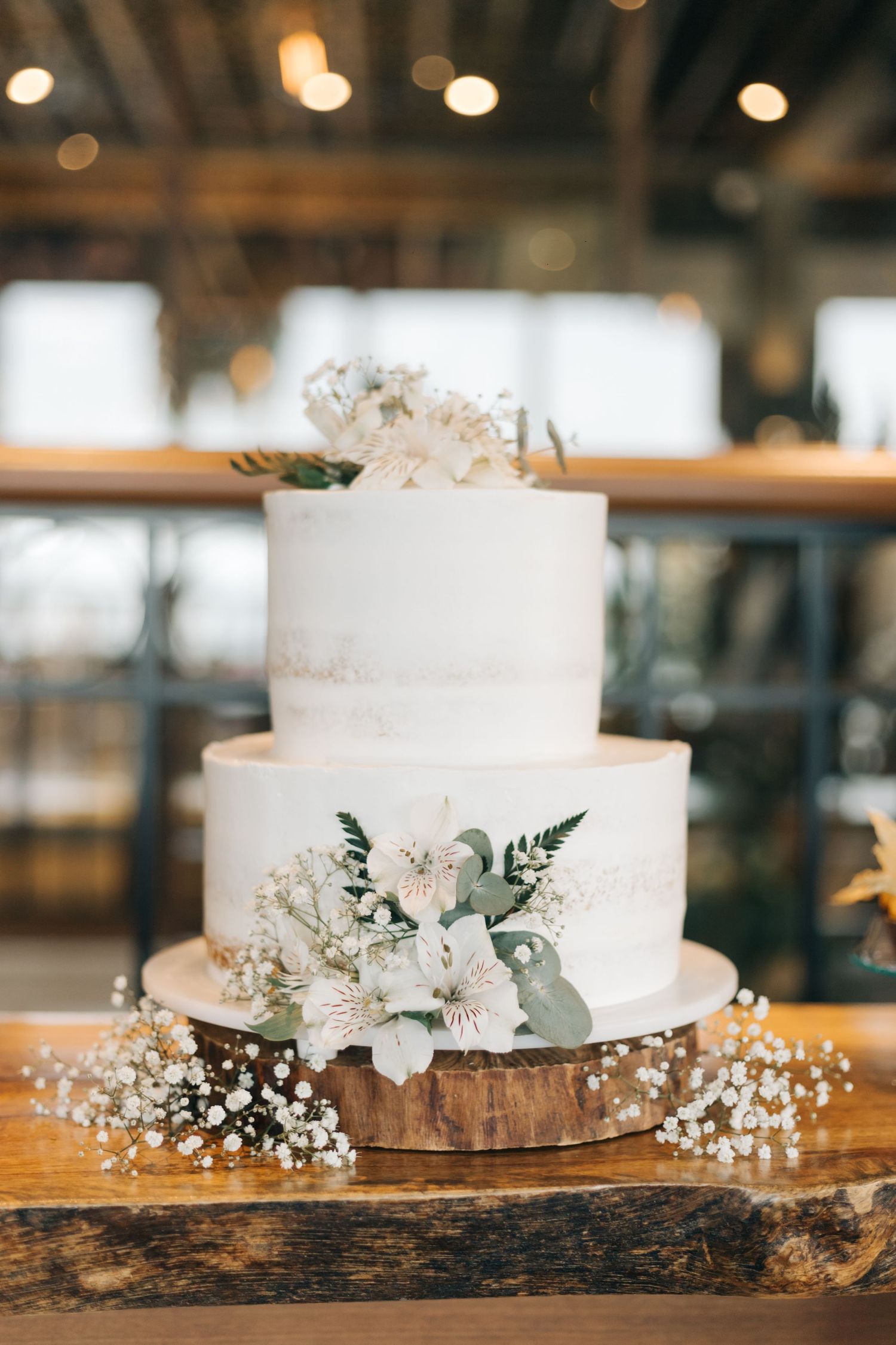 White wedding cake decorated with flowers and eucalyptus on rustic wood slice.