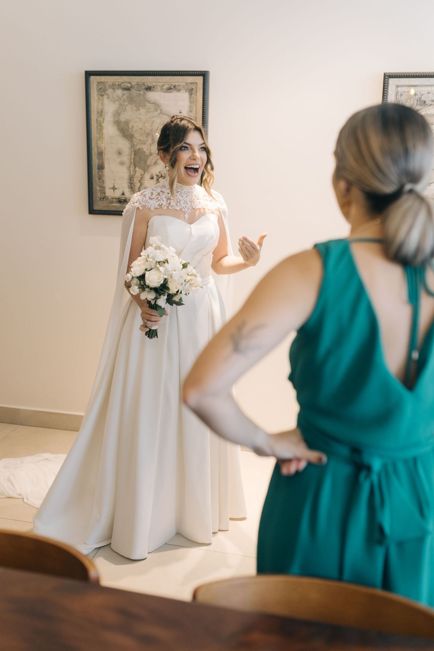 A bride in an elegant white gown laughs while holding a white bouquet near antique map artwork.