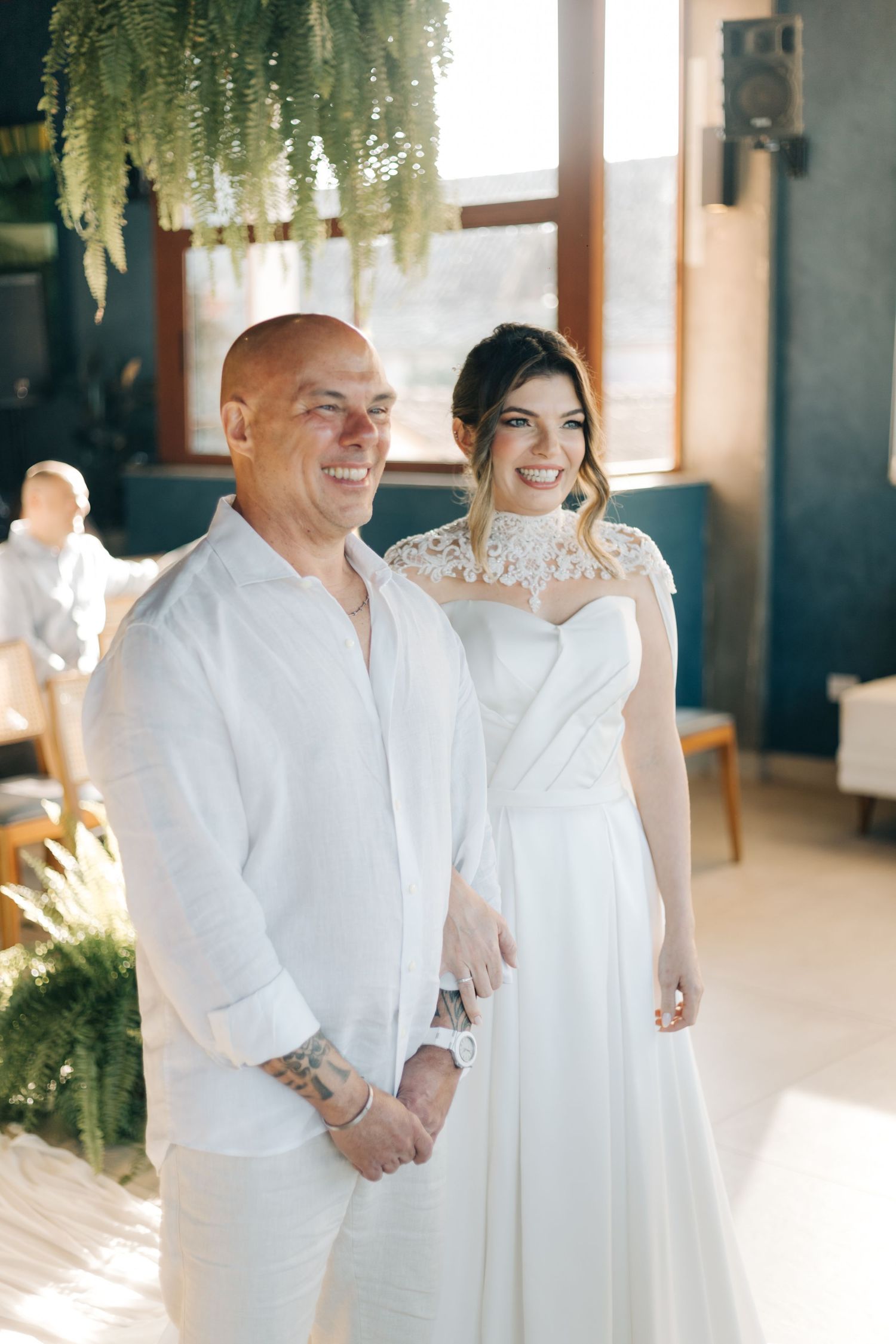 Couple in white wedding attire smiling together at an indoor venue with hanging floral decorations.