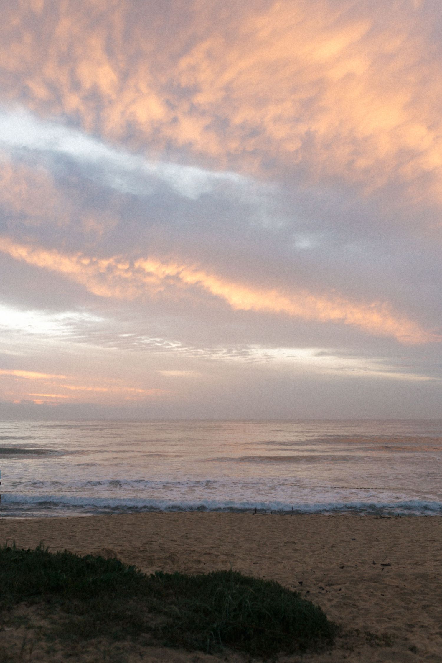 Panoramic coastal landscape at sunset with dramatic orange clouds reflecting over sandy beach and dark shrubs.