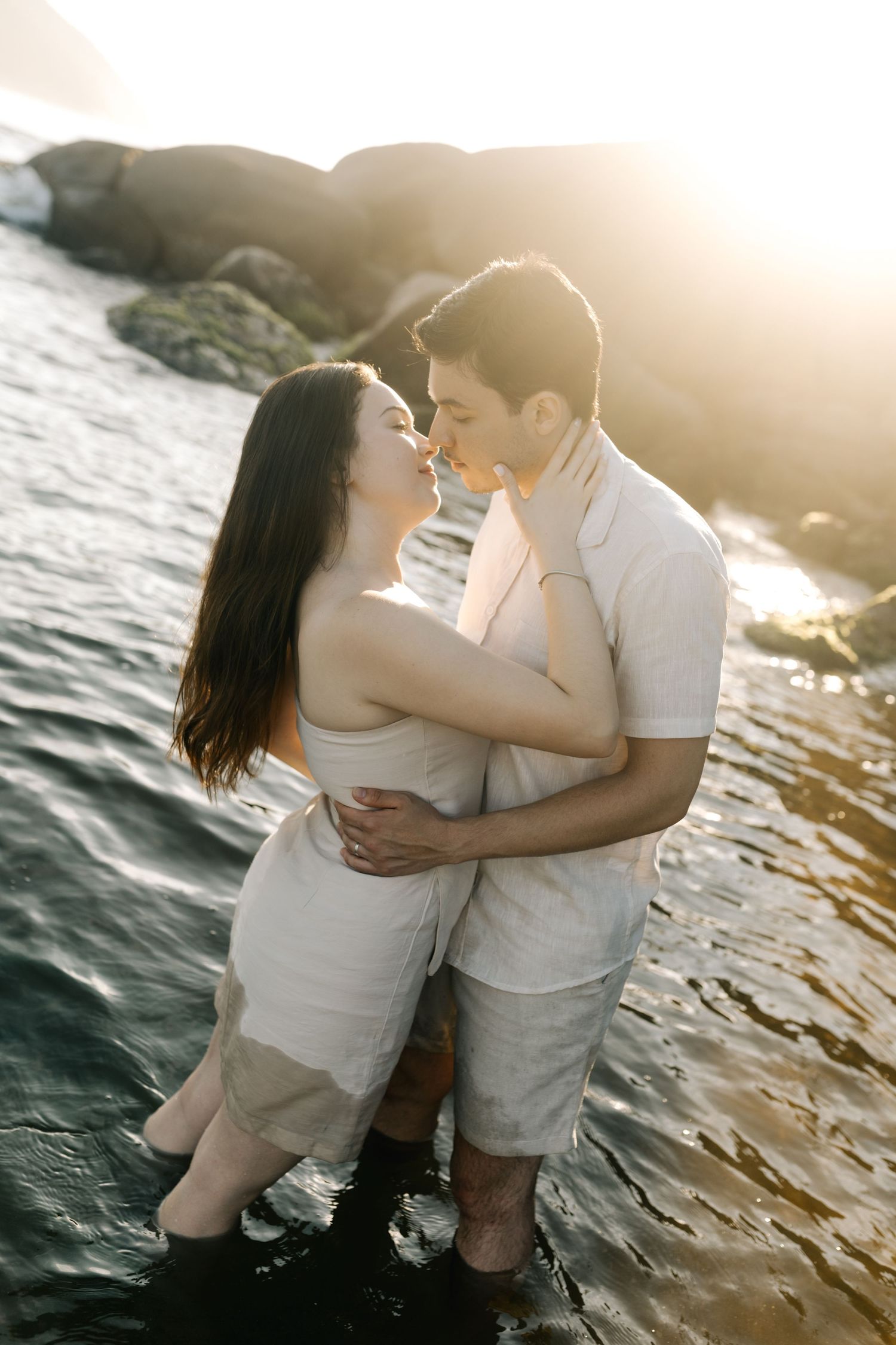 Couple embracing in water against sunset with rocks and golden light in background.