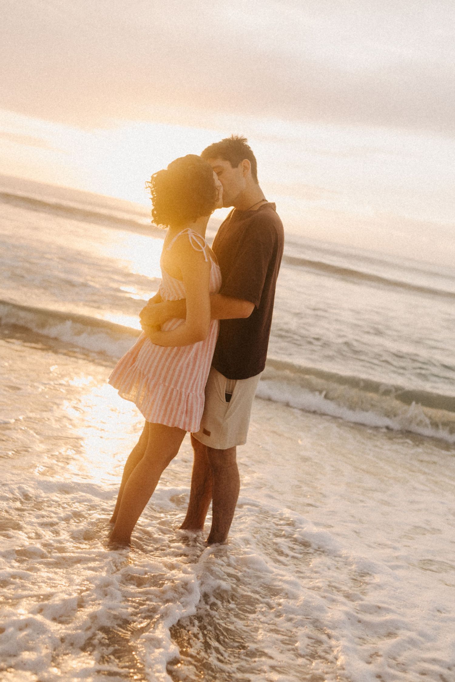 Um momento romântico ao pôr do sol na praia com um casal se abraçando nas ondas suaves do oceano enquanto a luz dourada ilumina a cena.