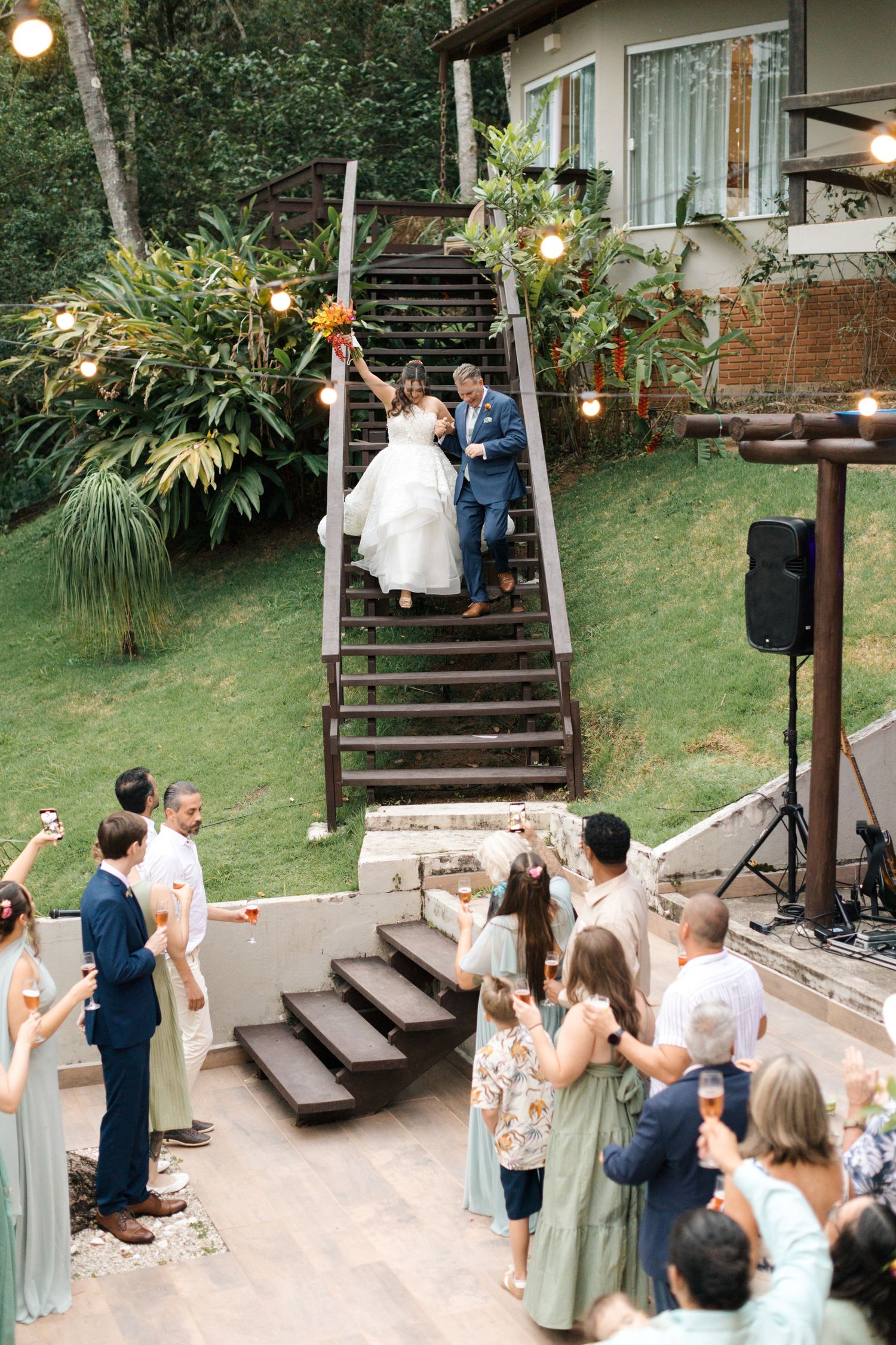 Wedding ceremony guests gathered on outdoor terrace with wooden steps and tropical landscaping.