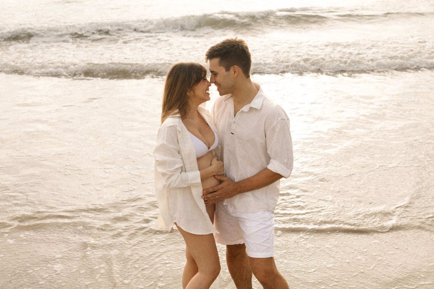 Romantic couple in white clothing embraces on sandy beach during golden hour sunset.