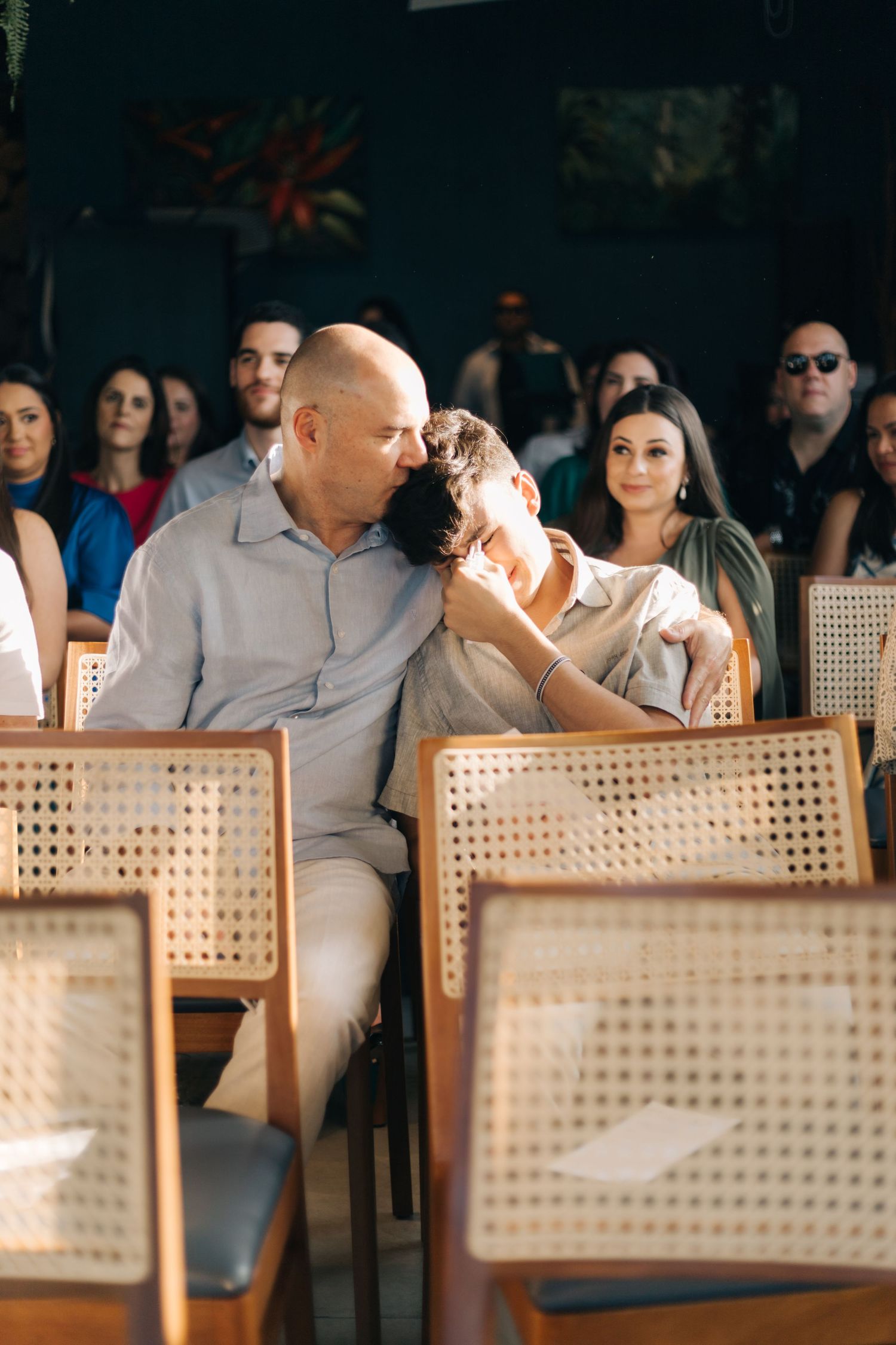 Wedding guests seated in cane-back chairs during indoor ceremony.