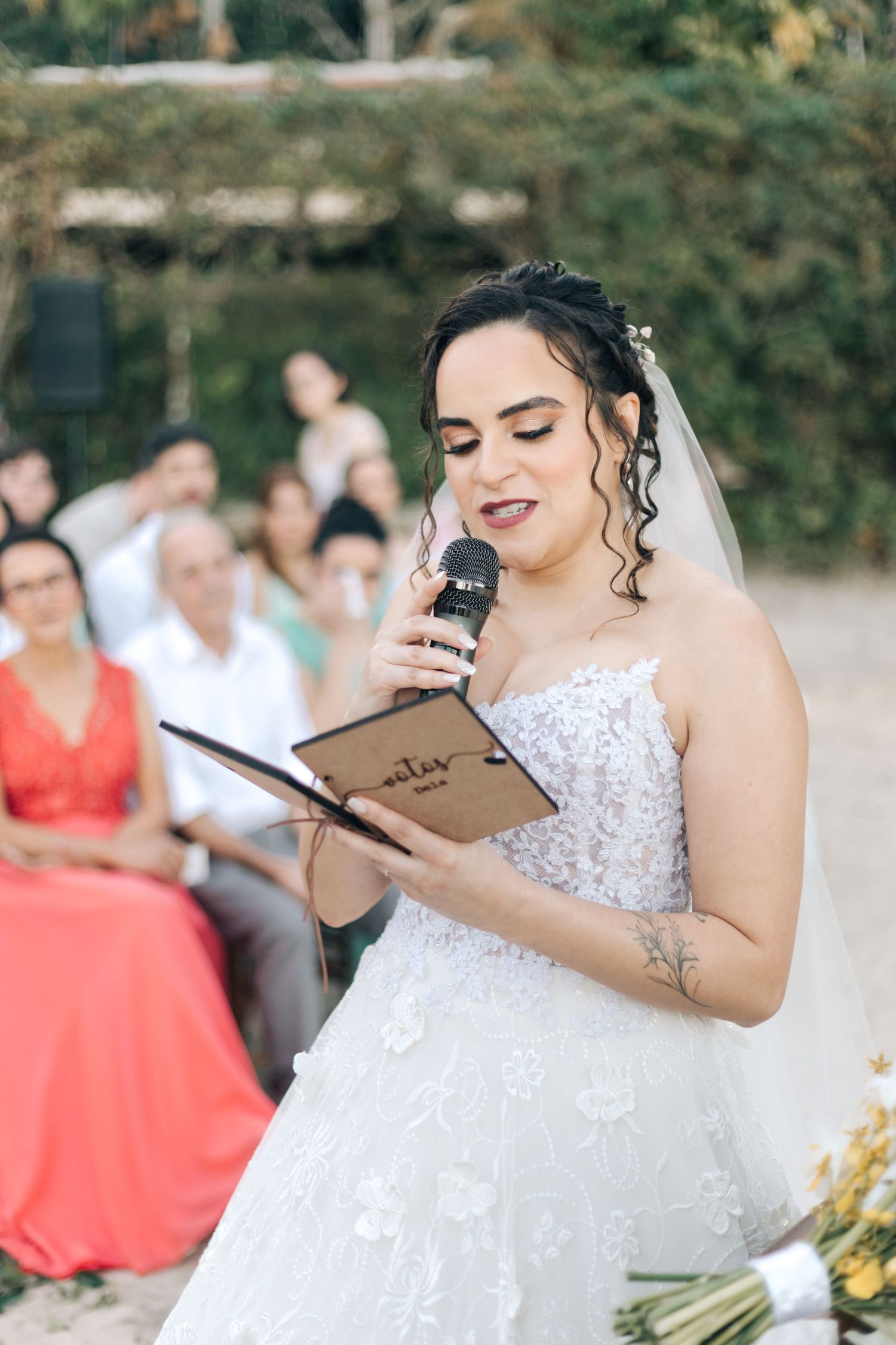 Bride in lace dress reads from a paper during emotional beach wedding ceremony.