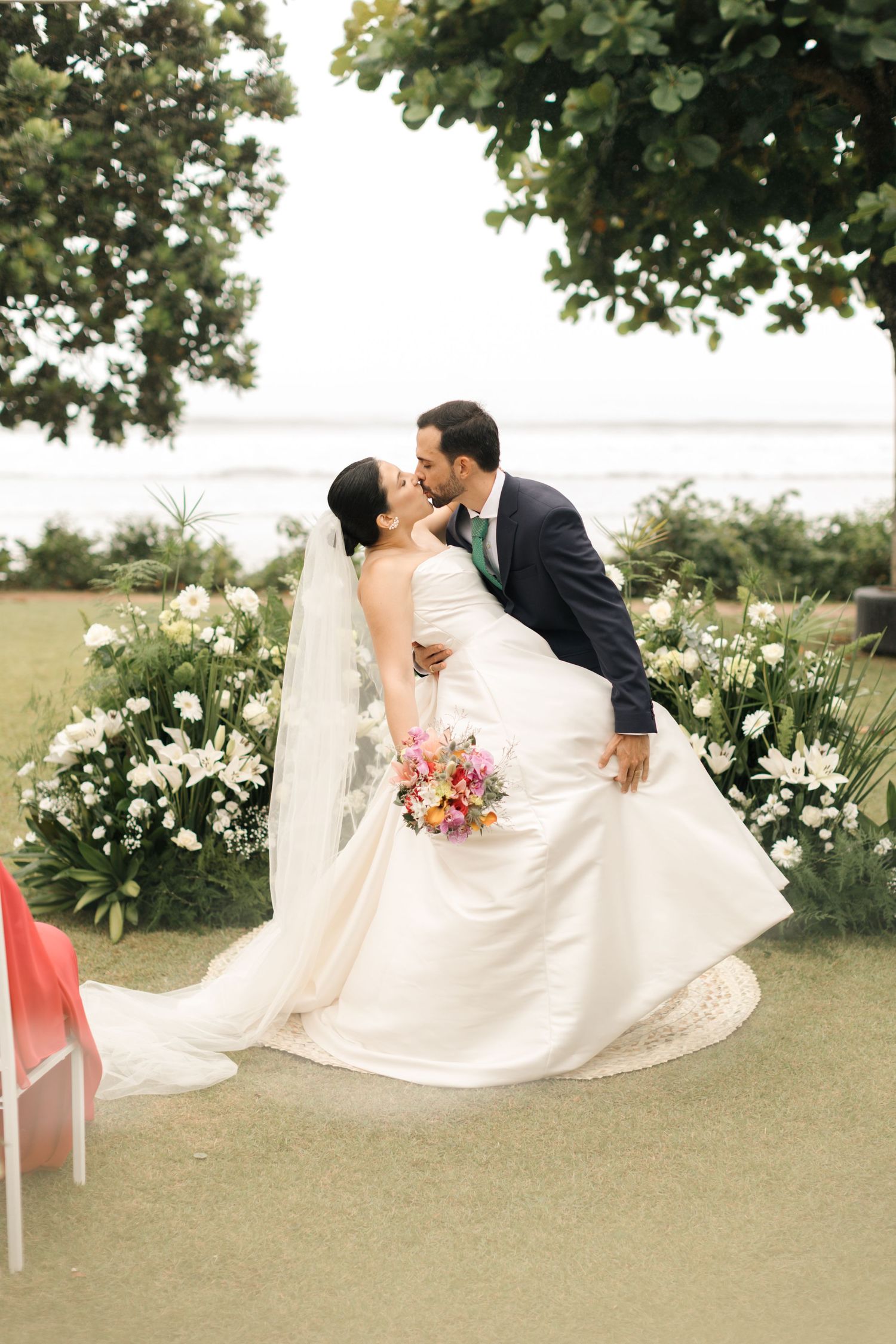 A romantic wedding portrait of a couple dipping and kissing among white flowers by the seaside.