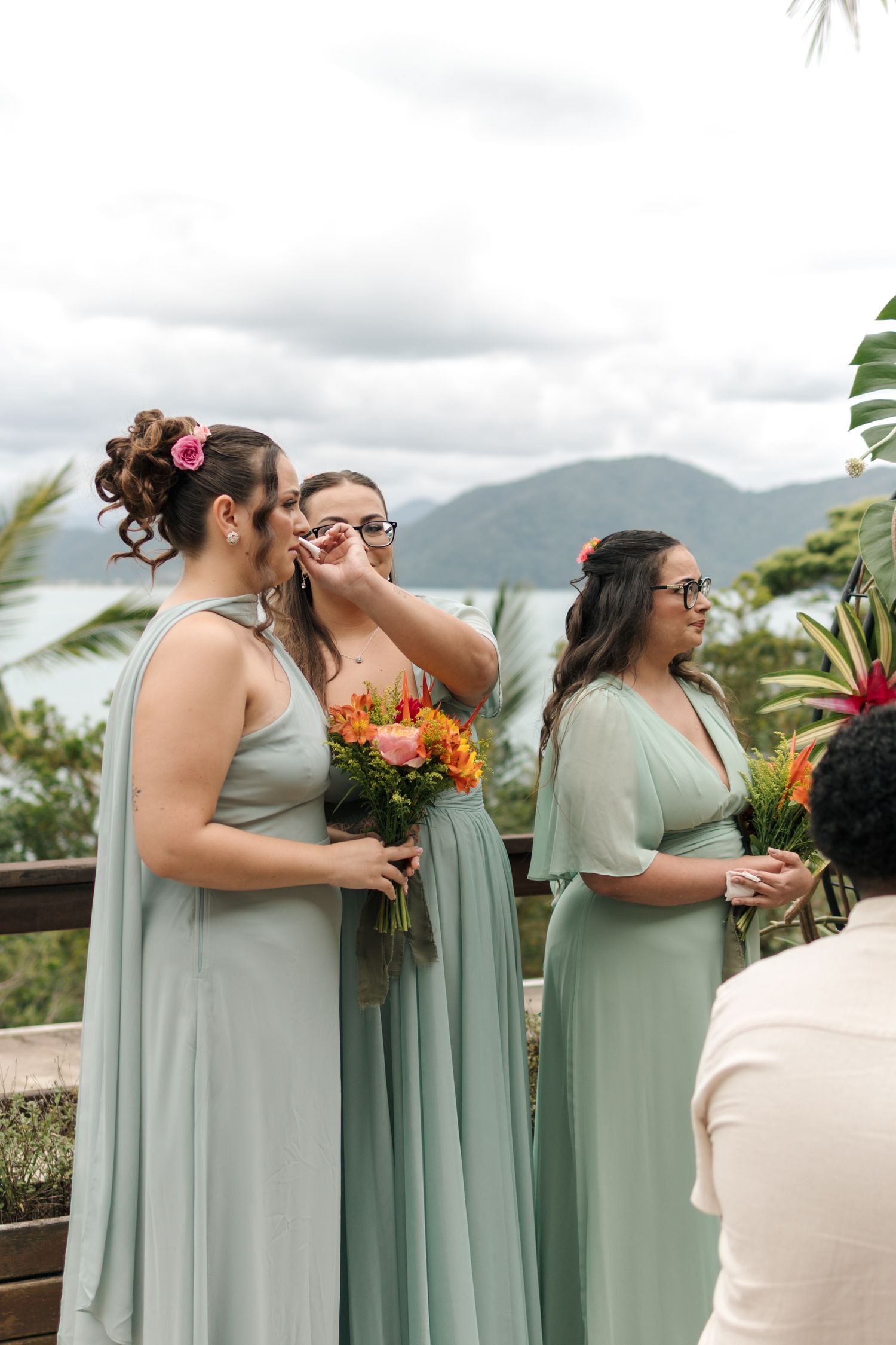 Bridesmaids in sage green dresses hold vibrant orange and pink bouquets against mountain backdrop.