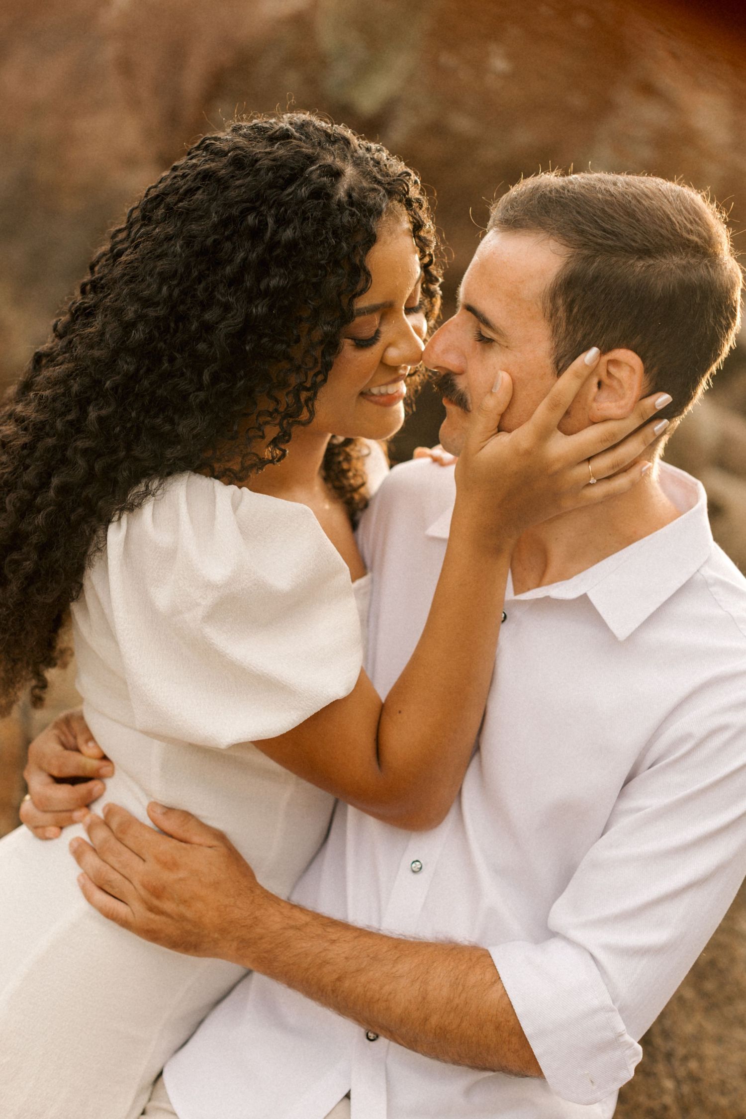 A romantic couple in white outfits share intimate close-up moments in a warm-toned outdoor setting.