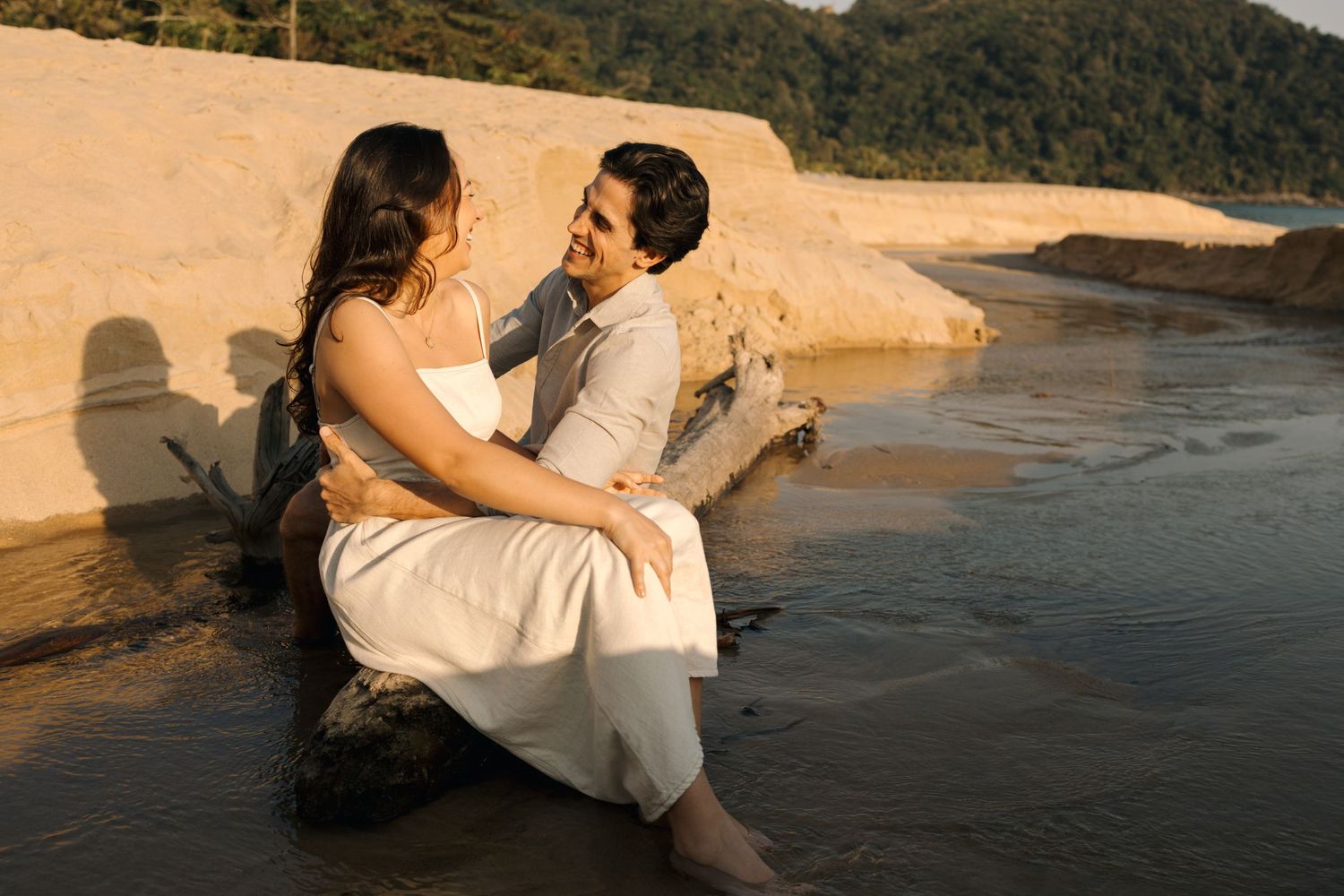 Romantic couple in wedding attire embraces on sunlit beach at golden hour.