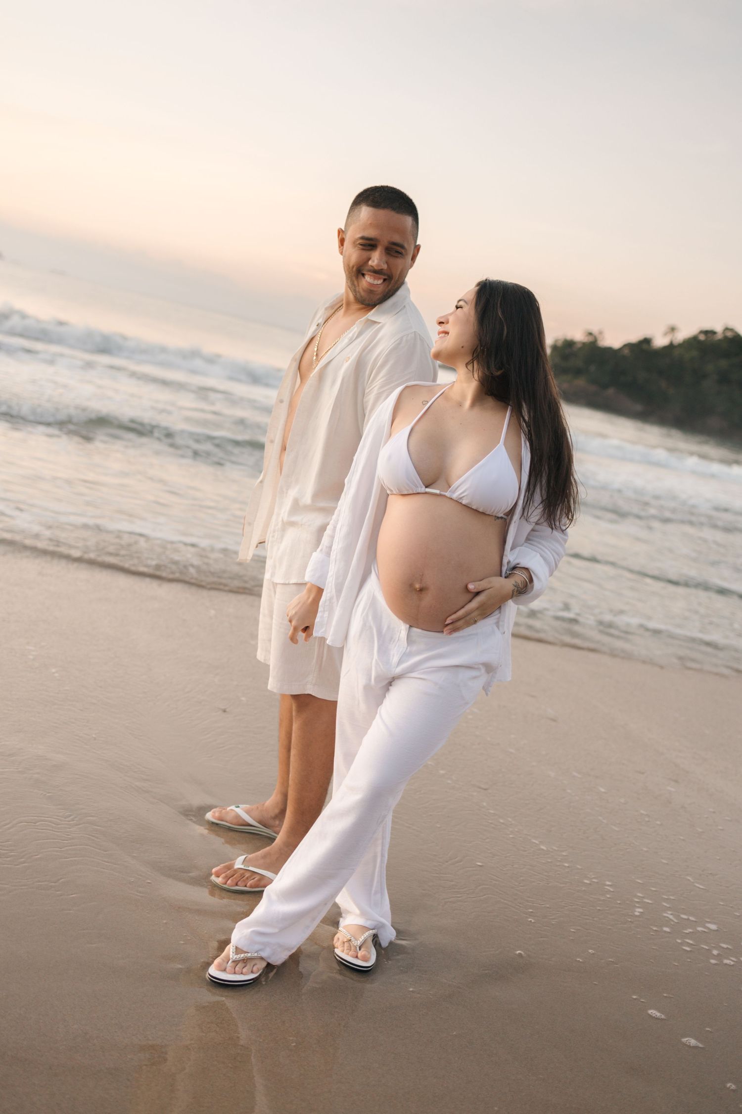 Joyful couple in white outfits posing for maternity photos on a beach at sunset.