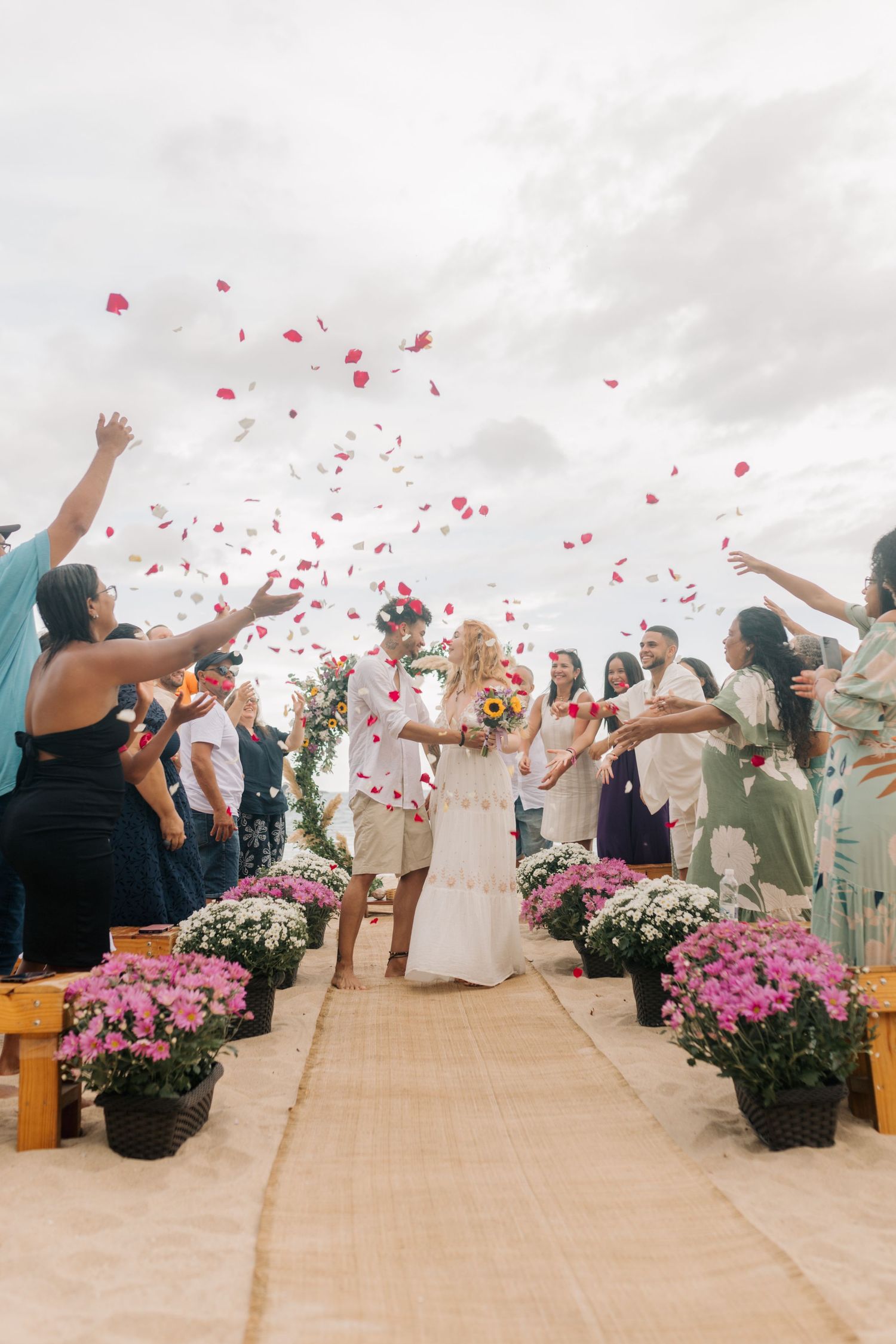 Wedding guests toss rose petals as newlyweds walk down aisle lined with pink flowers on beach.