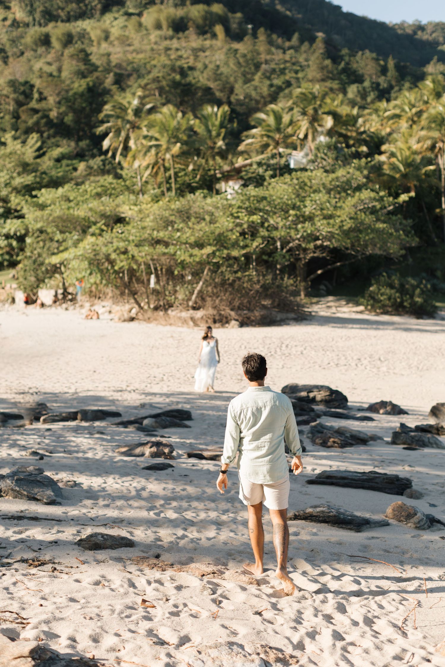 Person walking on a scenic tropical beach with palm trees and rocks along the shoreline at sunset.