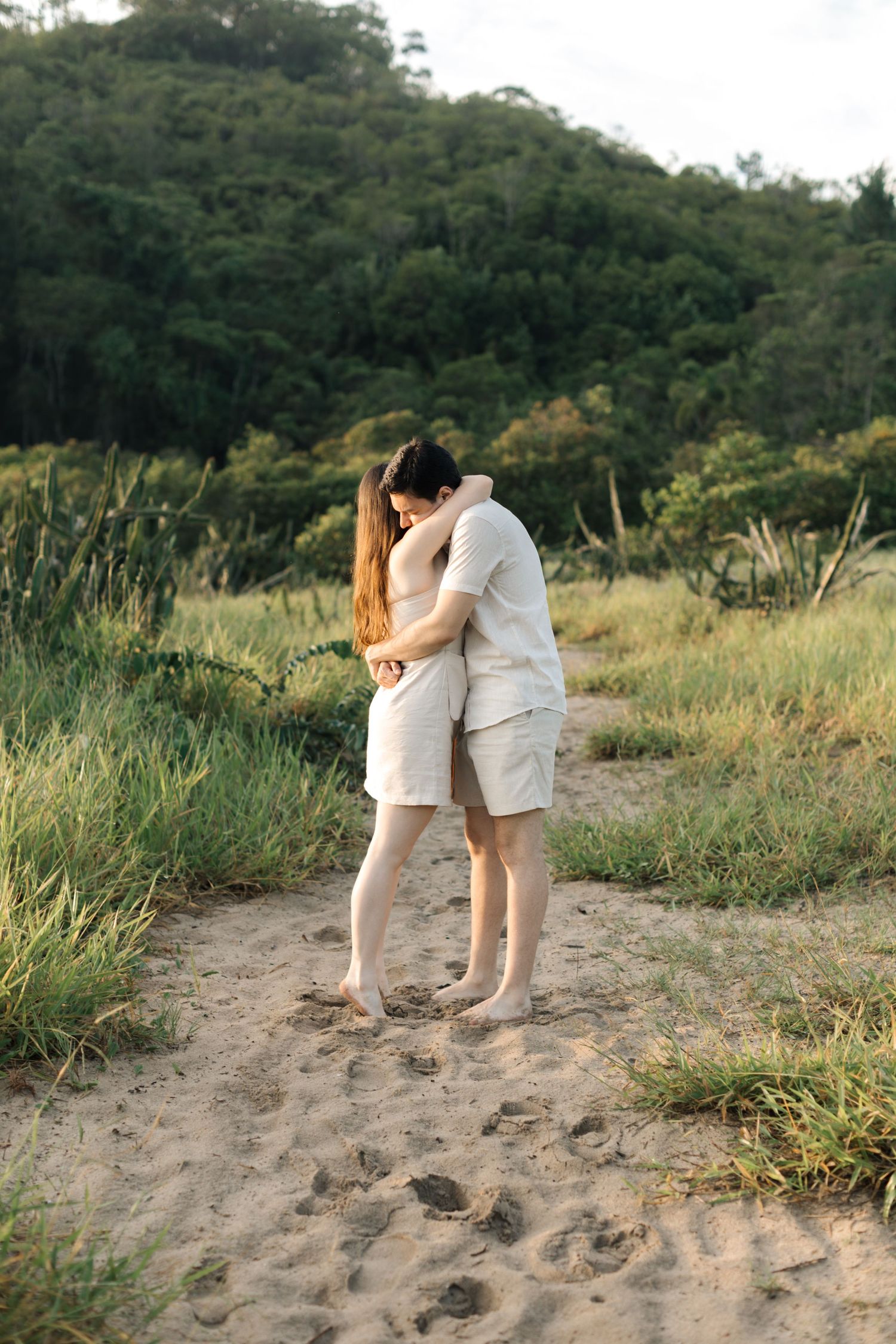 Couple sharing intimate moment on sandy trail with lush green hills in background.