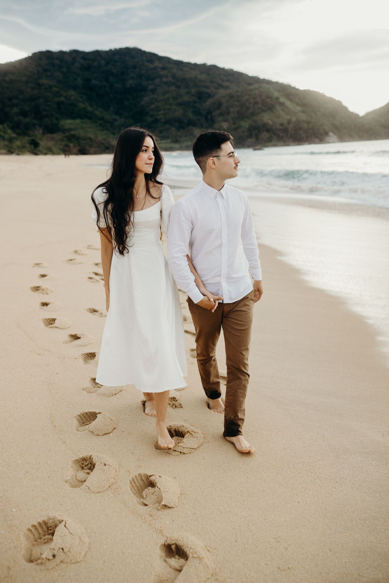 Couple in white outfits walking barefoot on a sandy beach leaving footprints behind them.