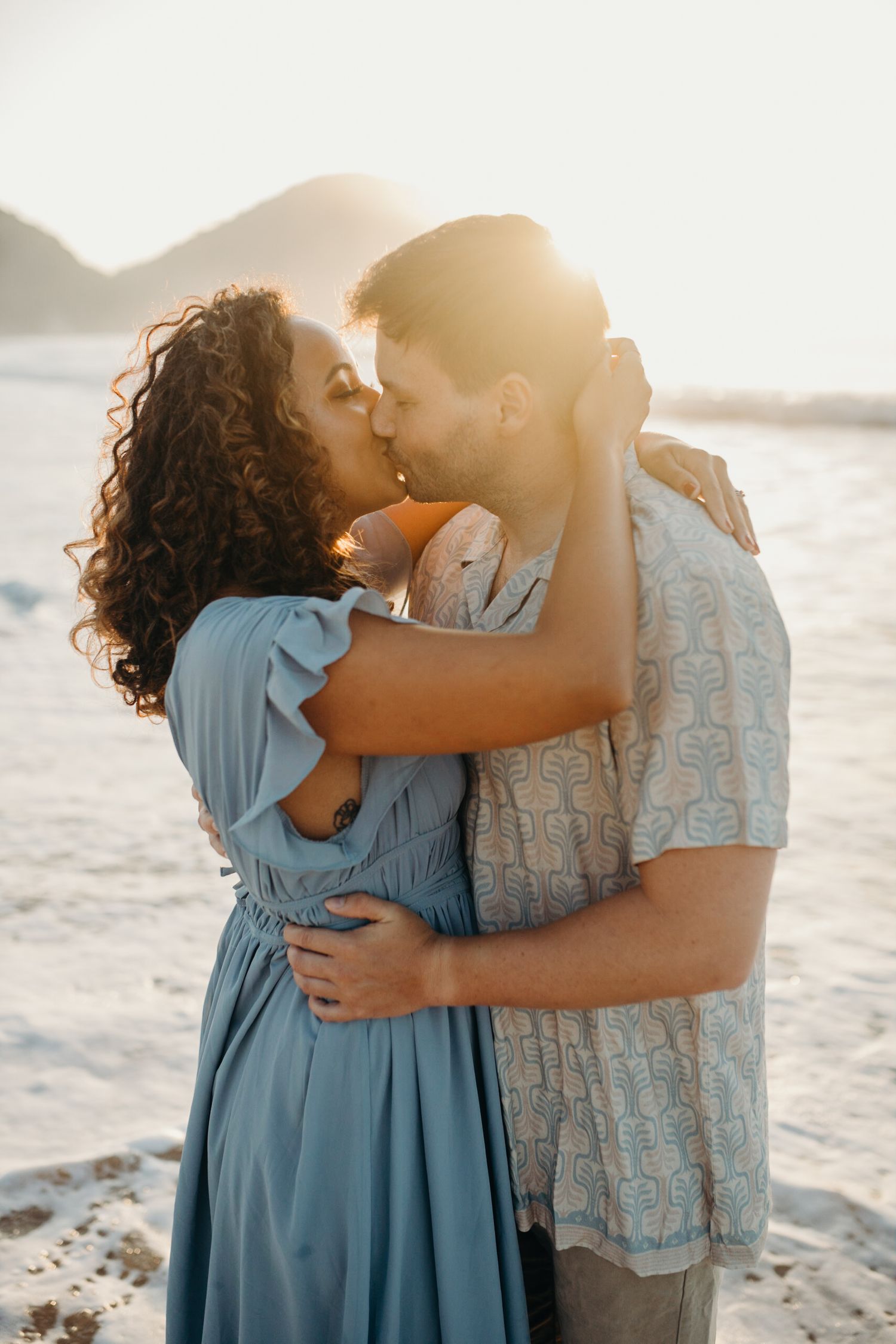 Romantic beach photoshoot with couple in blue dress and patterned shirt.