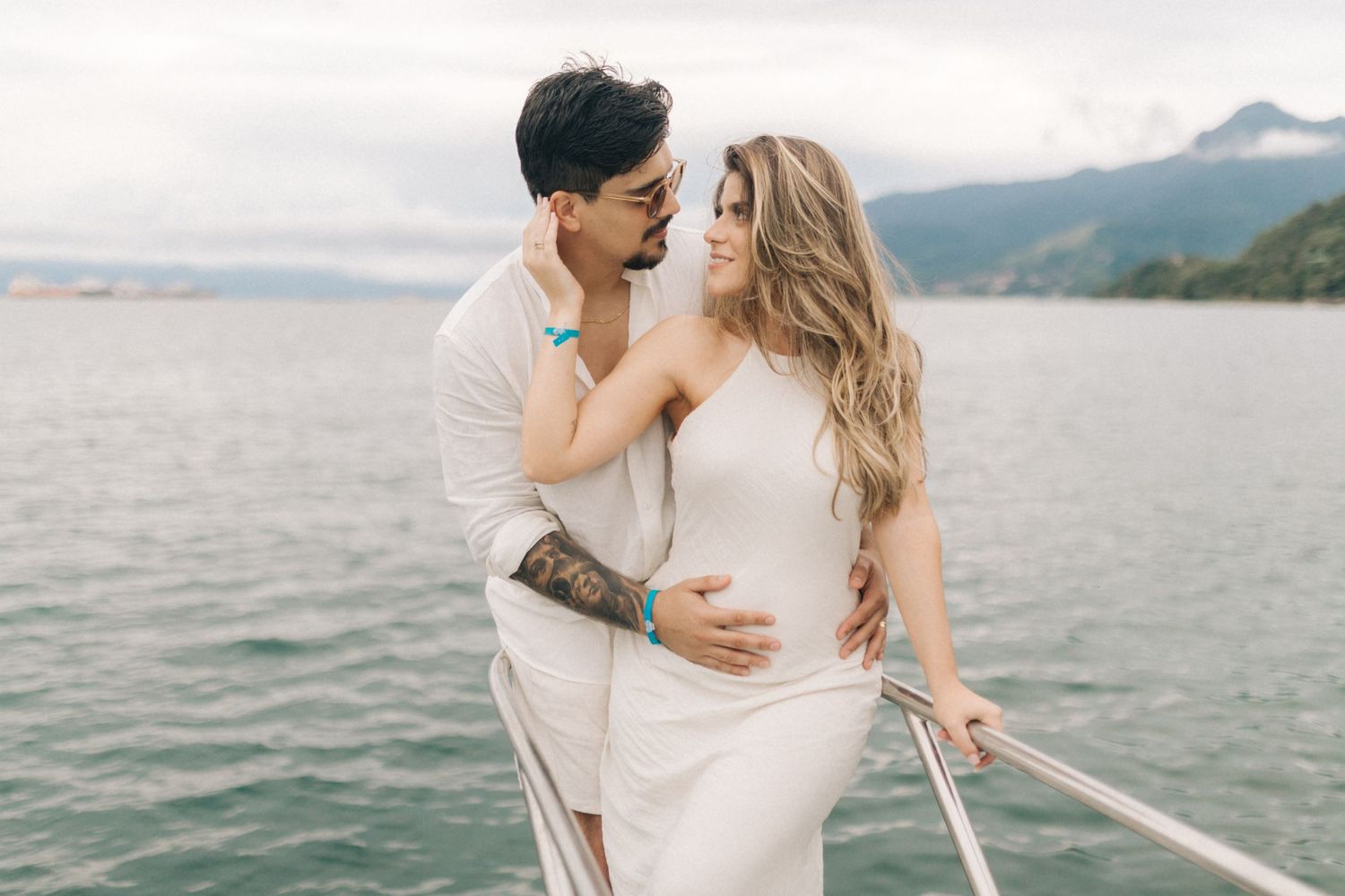 A romantic couple in white outfits embracing on a boat with mountains and water in the background.