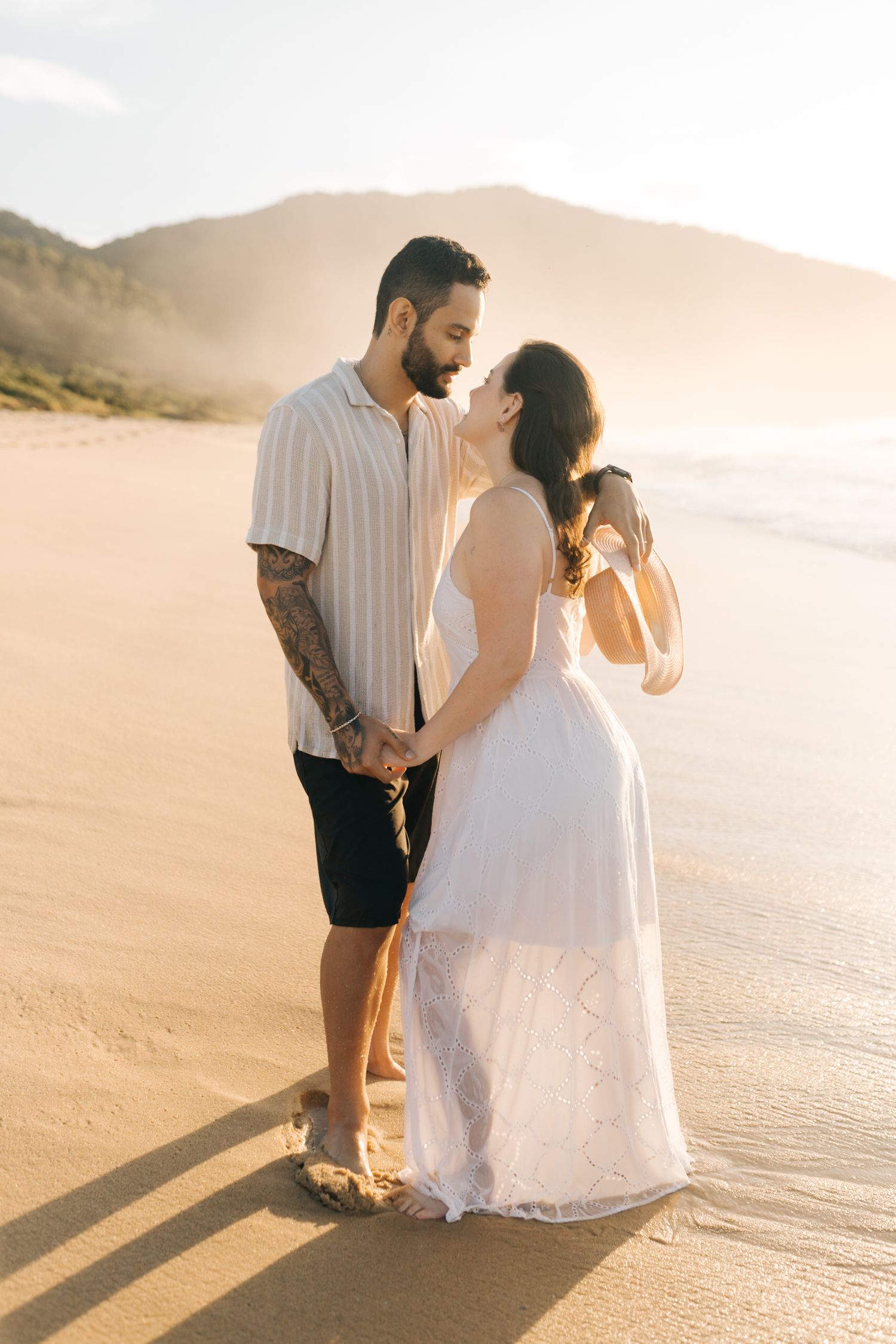 A romantic couple walks together on a sandy beach during golden hour.