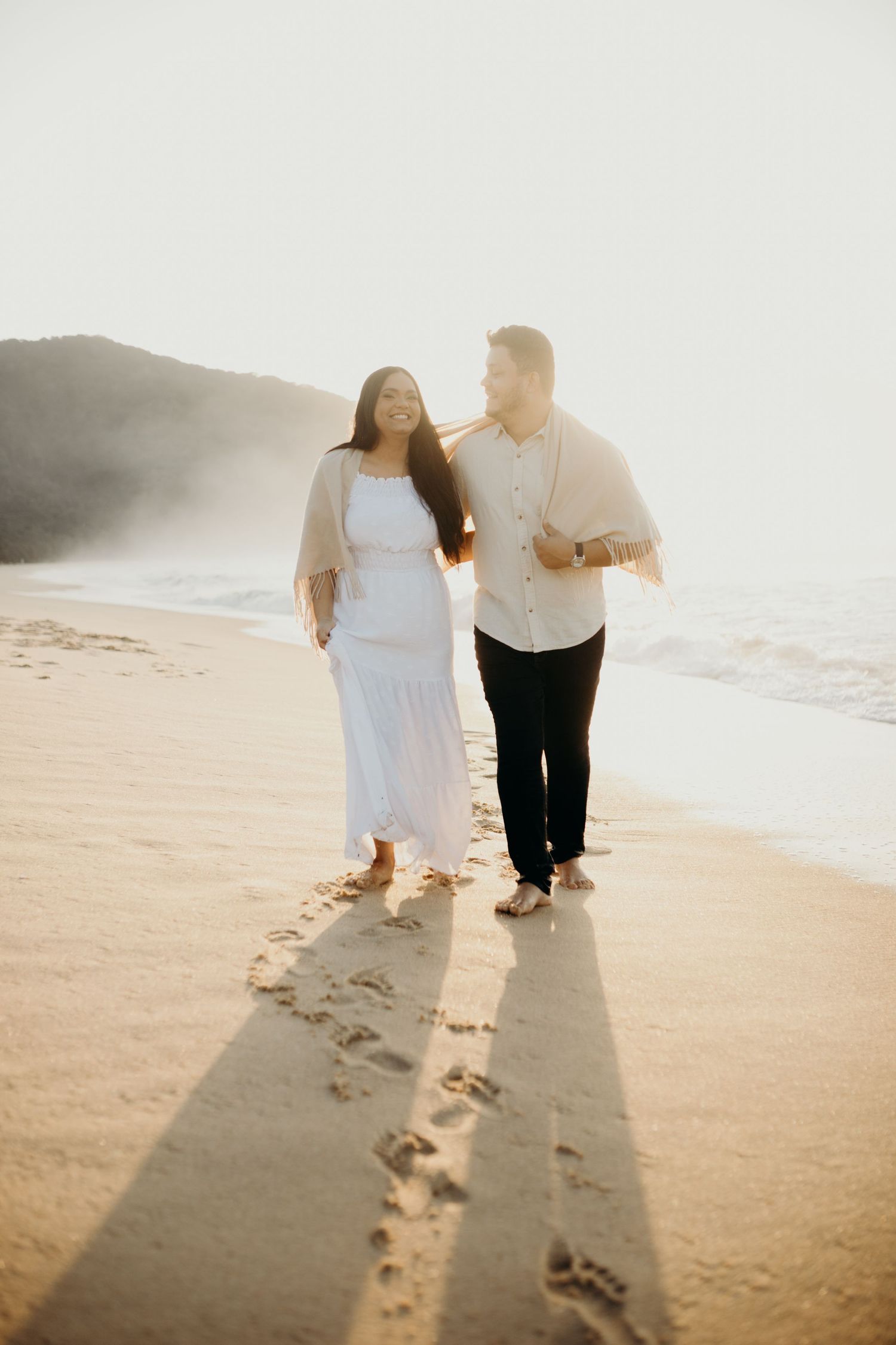 Long shadows cast on beach sand as couple walks together during sunset.