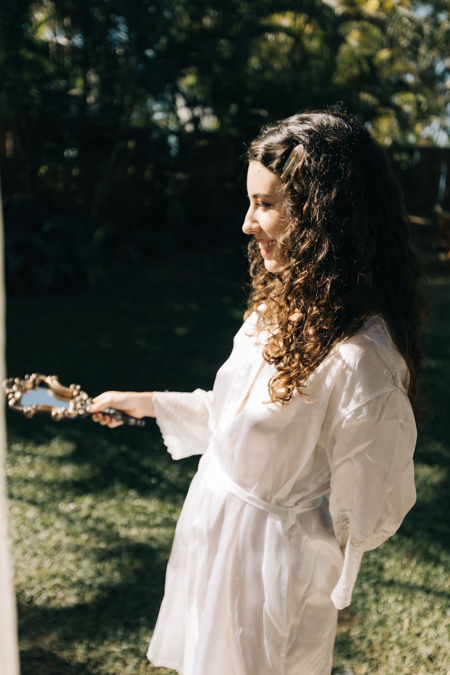 Person in flowing white dress with curly hair standing in sunlit garden.