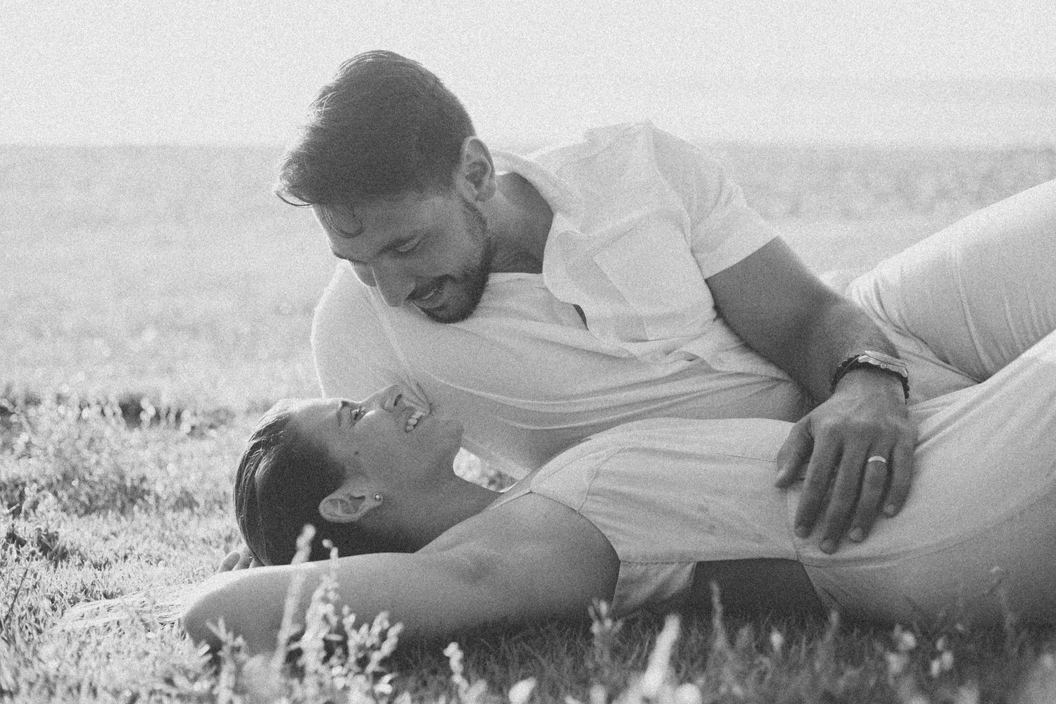 Black and white photo of couple lying in grass field during romantic photoshoot.