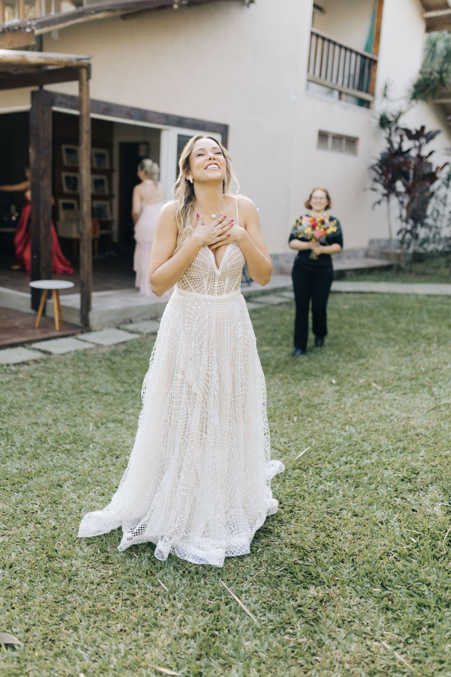 A beaming figure in a flowing white wedding dress stands on a lawn near a building.