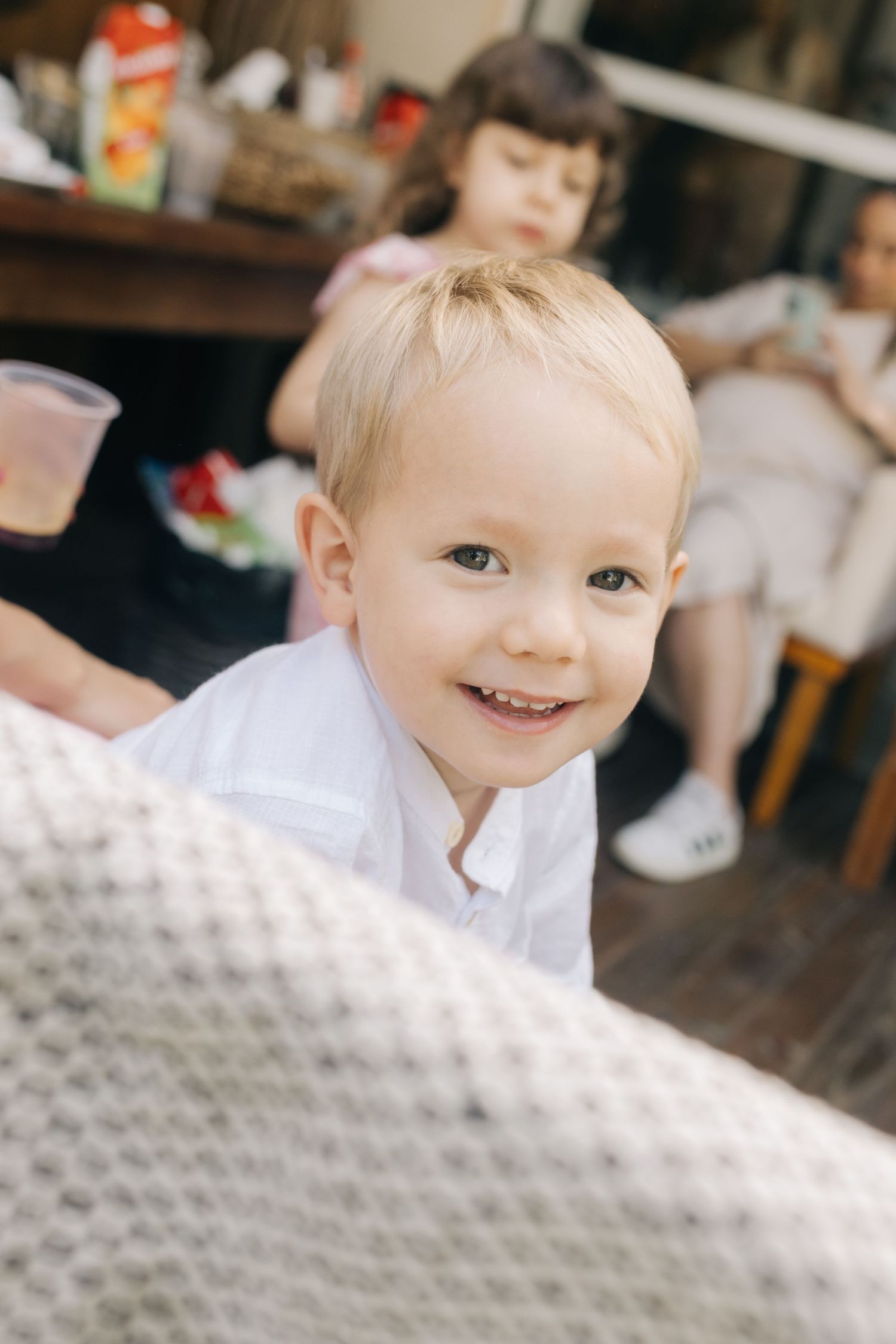 A young child in a white shirt smiles brightly at the camera during an indoor gathering.