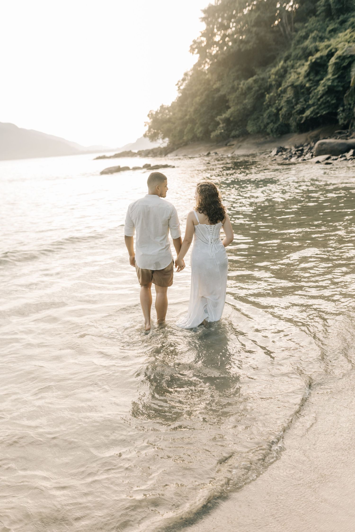 Couple walking hand in hand through ocean waves during golden hour.