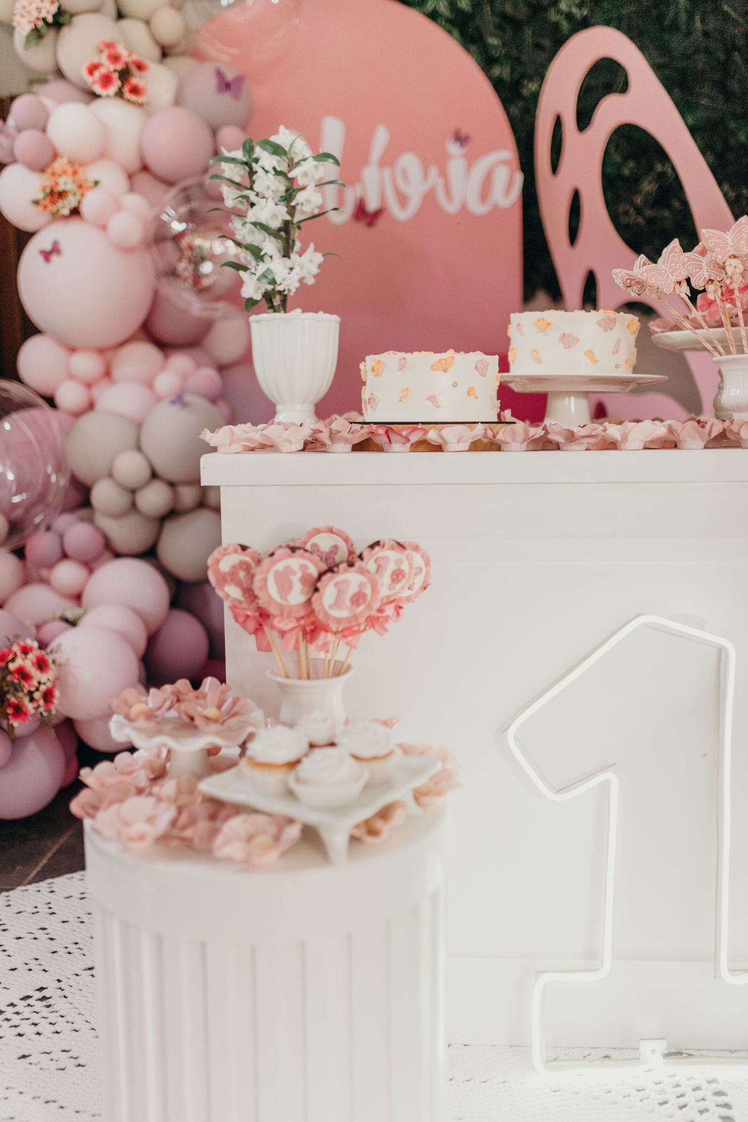 Pink and white first birthday party setup with balloon garland and dessert displays.