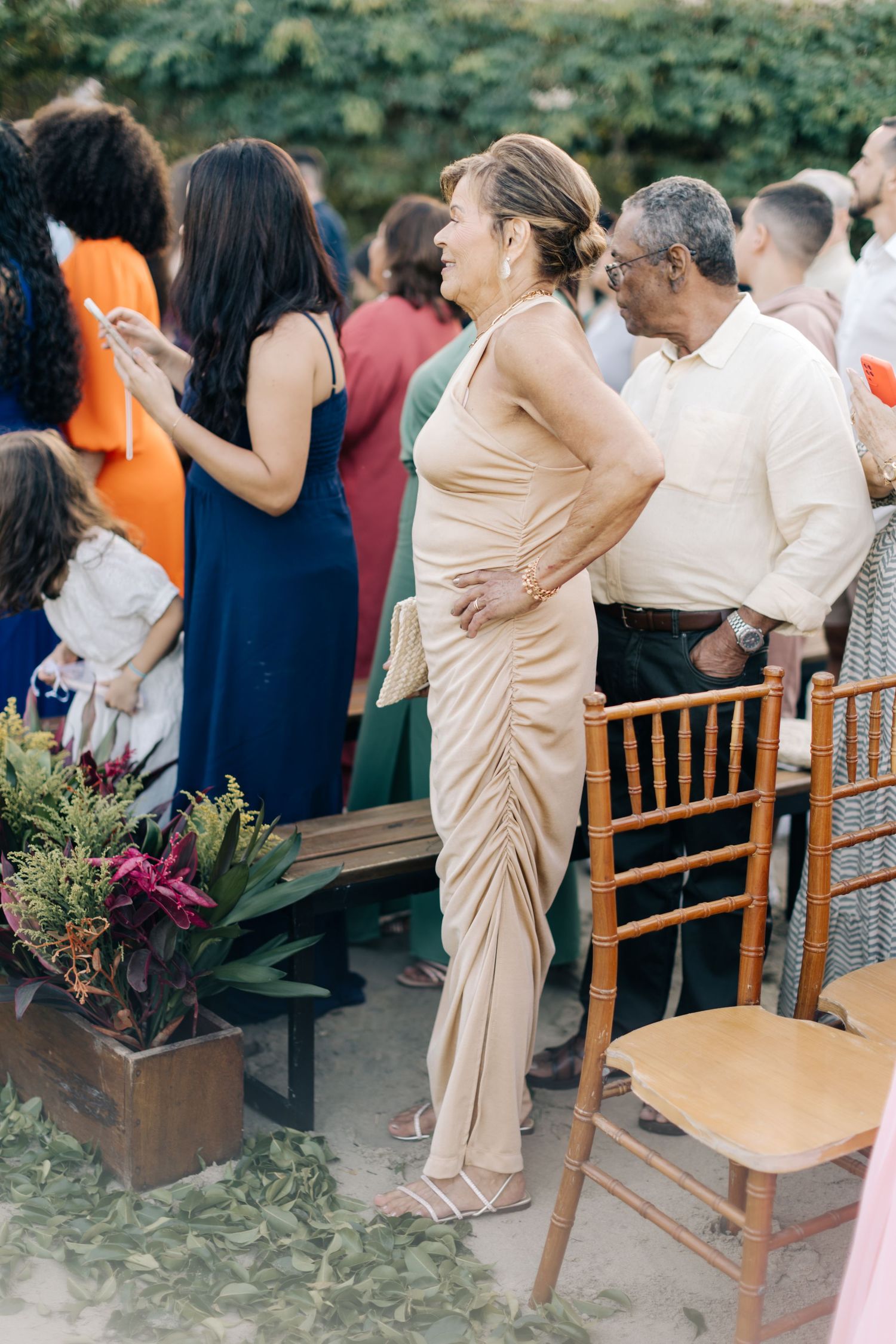 Guests gather at outdoor event with wooden chairs and tropical floral decorations.