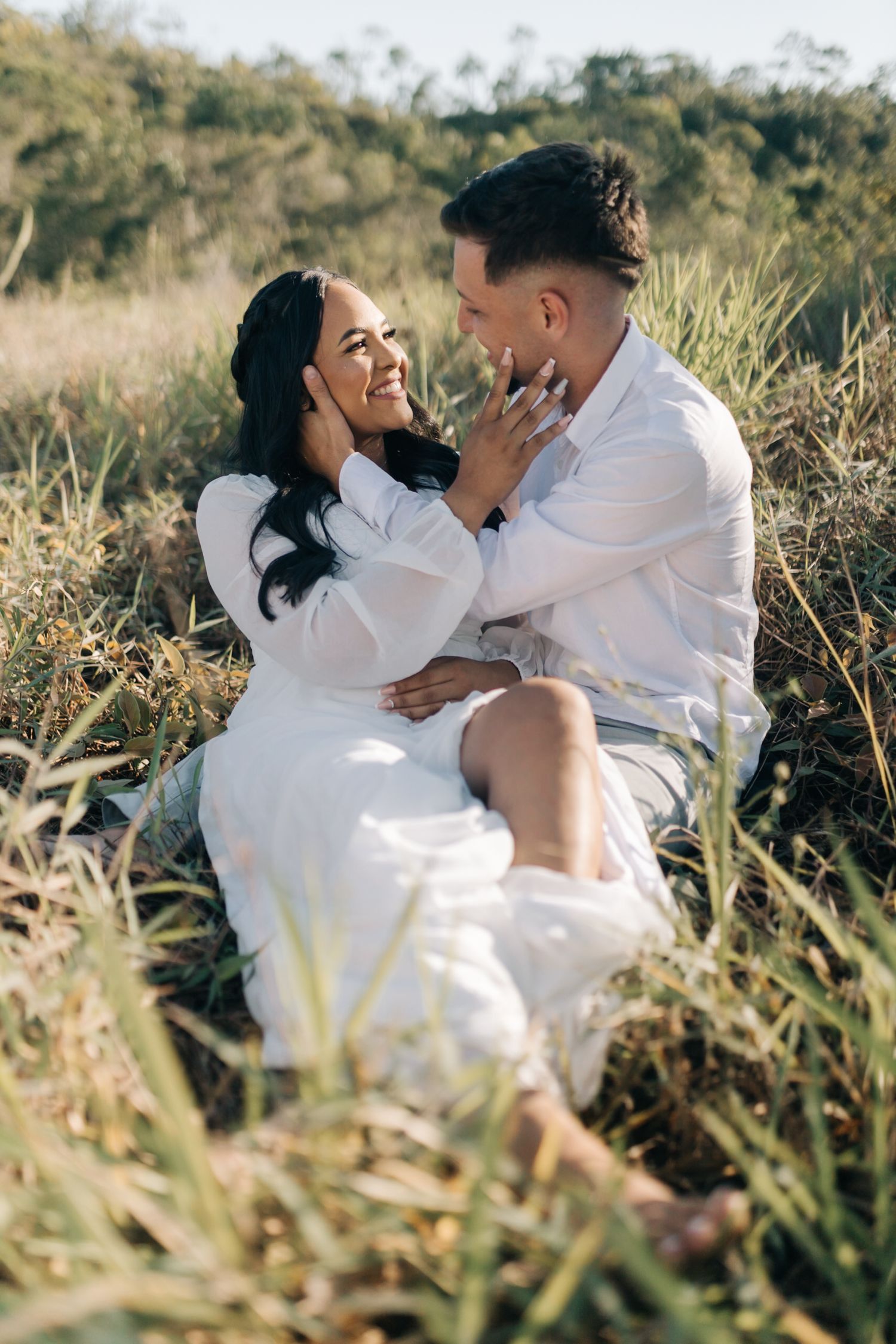 A couple in white clothing shares an intimate moment while sitting in tall grass at sunset.