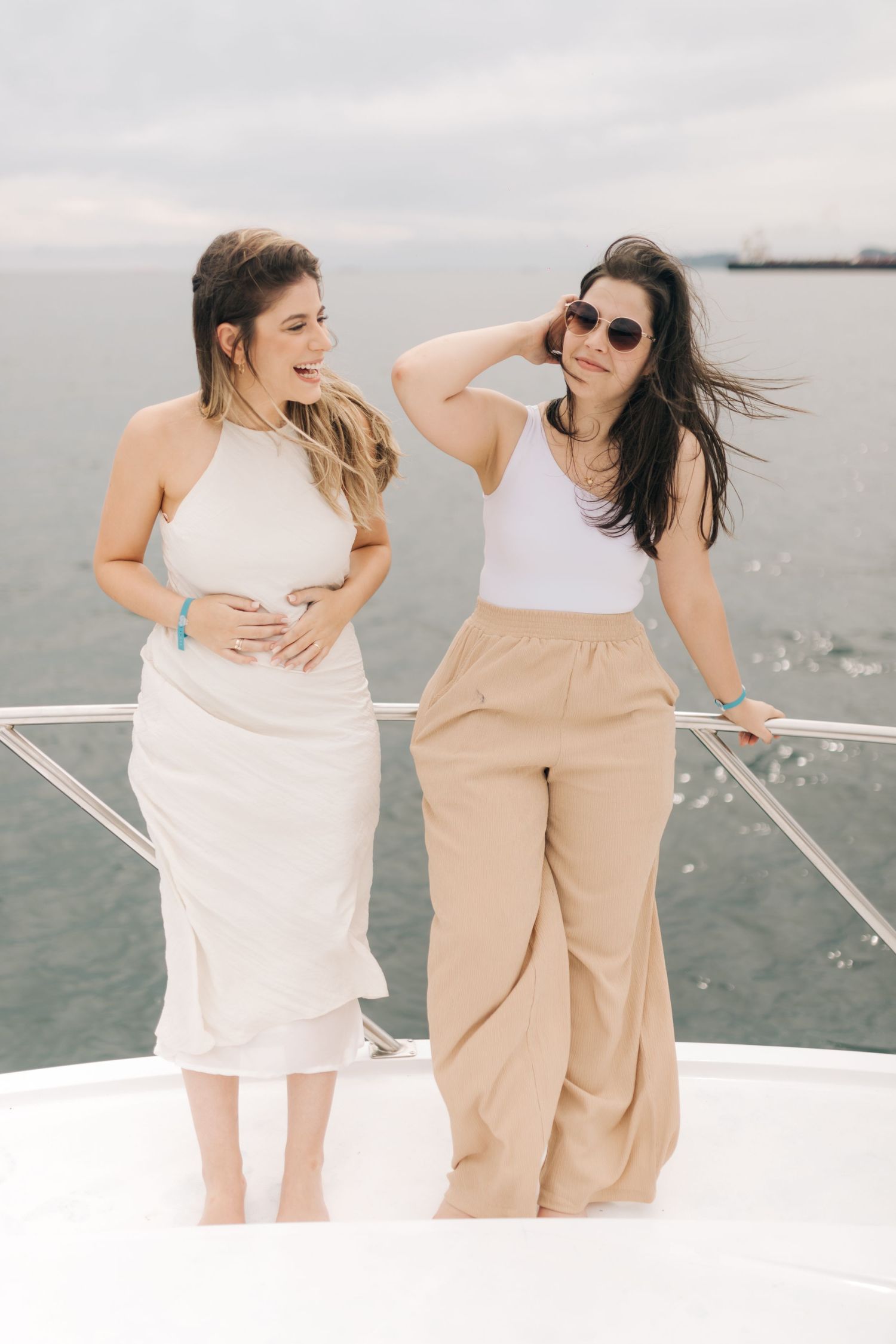 Two friends in summer outfits laughing together on a boat deck with ocean and cloudy sky in background.