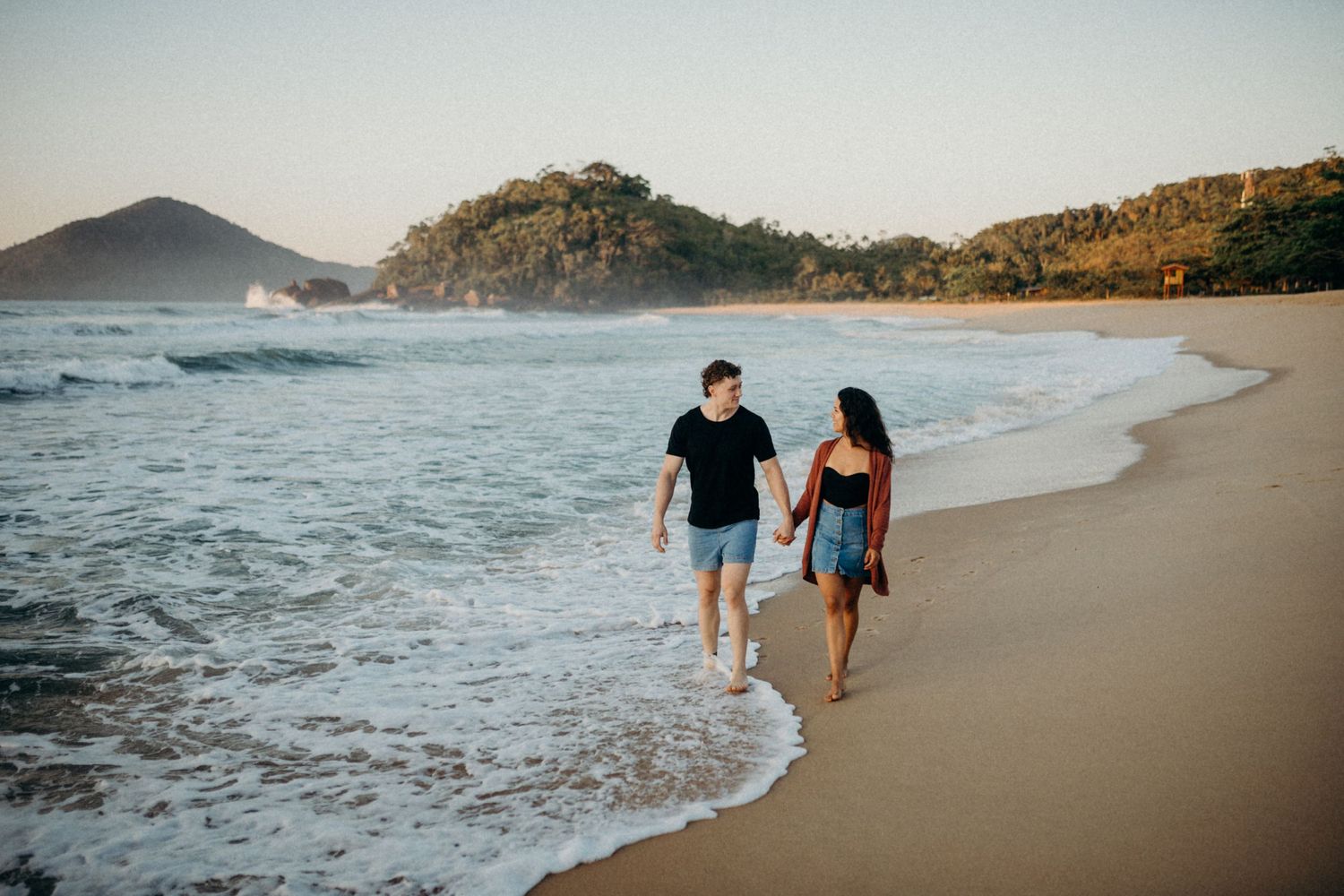 Couple walking along beach shoreline with mountains and waves in background.