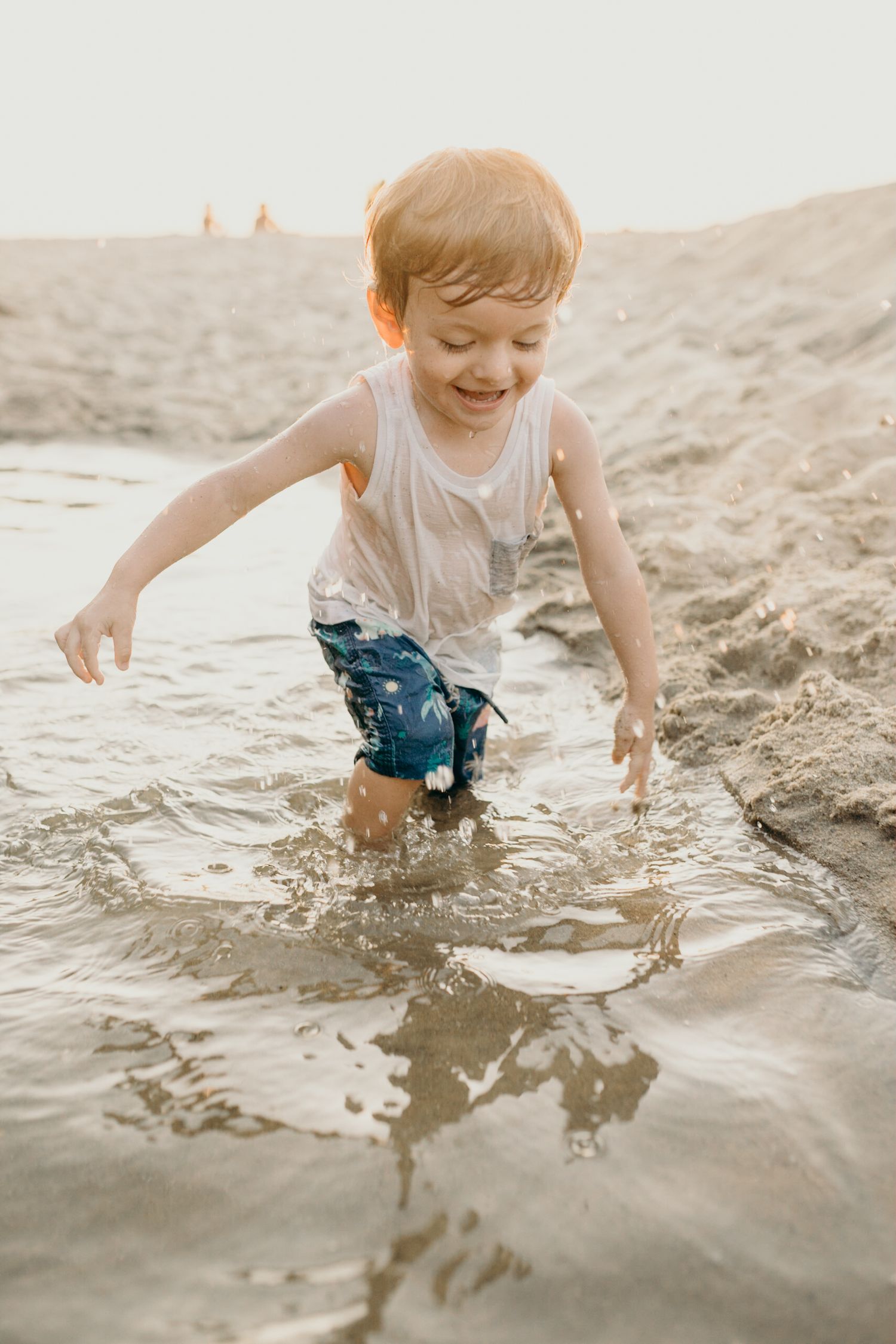 Joyful child splashes and plays in shallow beach water wearing a white tank top.