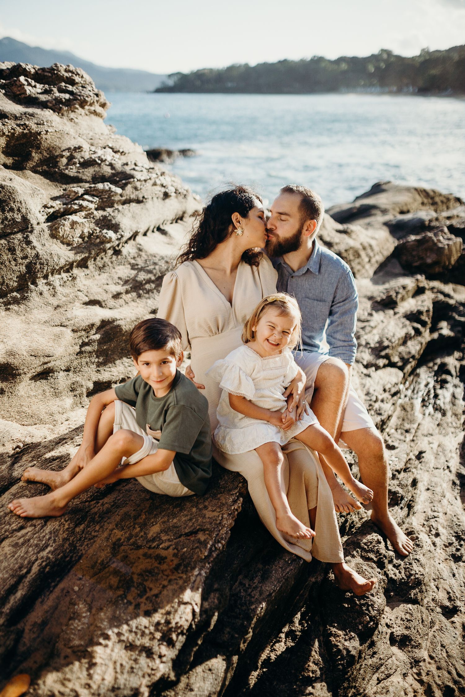 A loving family of four sits together on rocky coastal terrain with the ocean in the background.