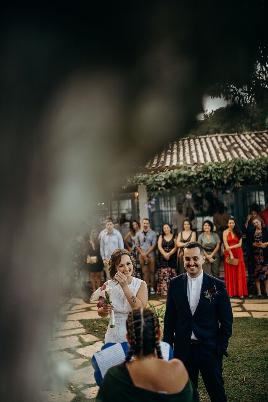Guests gather at an outdoor wedding ceremony with a rustic building's tiled roof visible in the background.