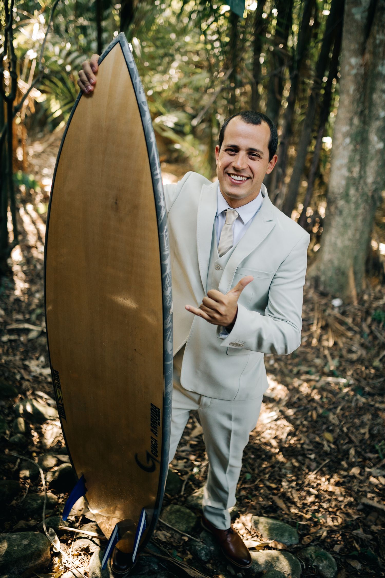 A person in a light colored suit poses with a surfboard in a wooded setting.