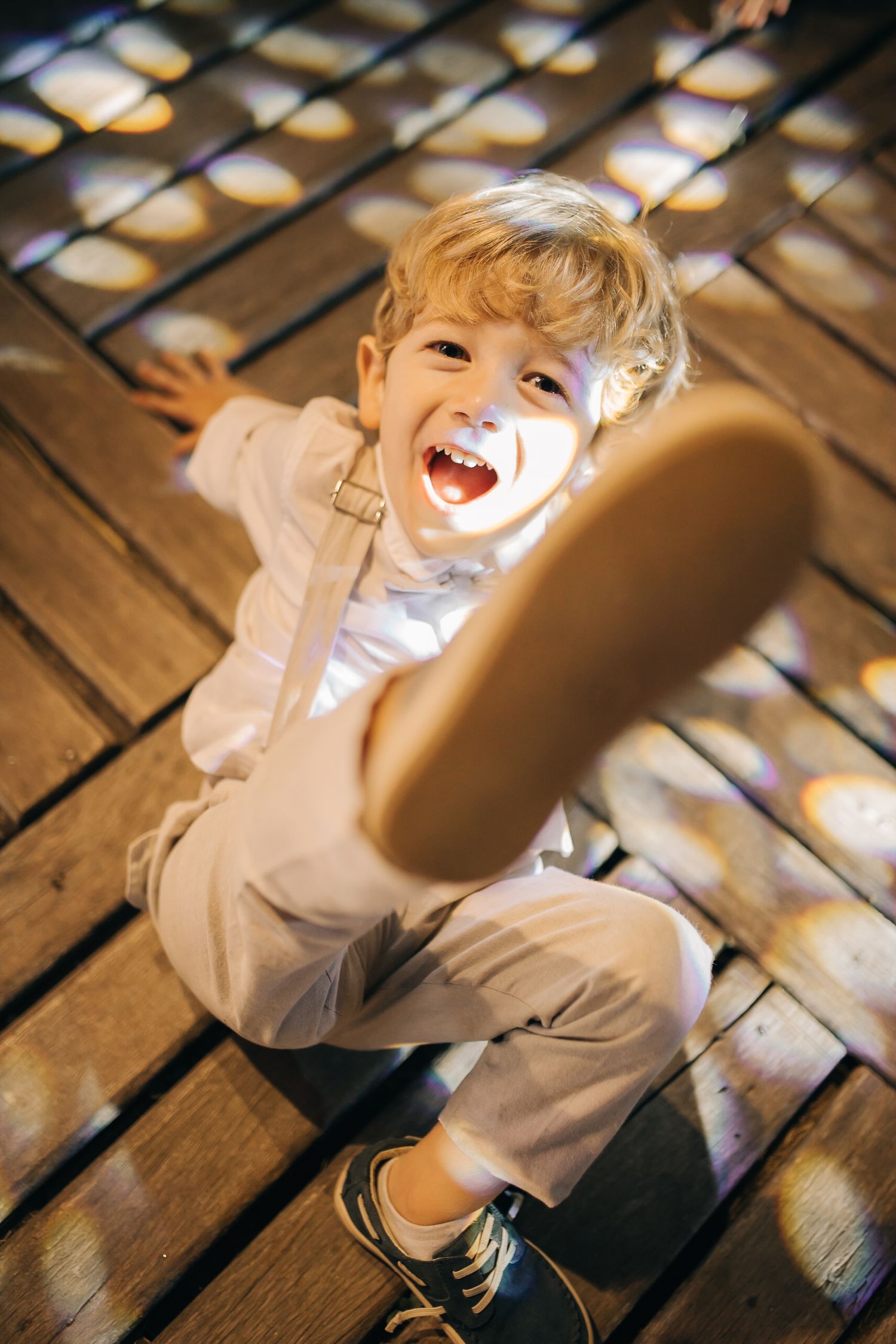 Young child in beige suit laughs joyfully while sitting on wooden deck.