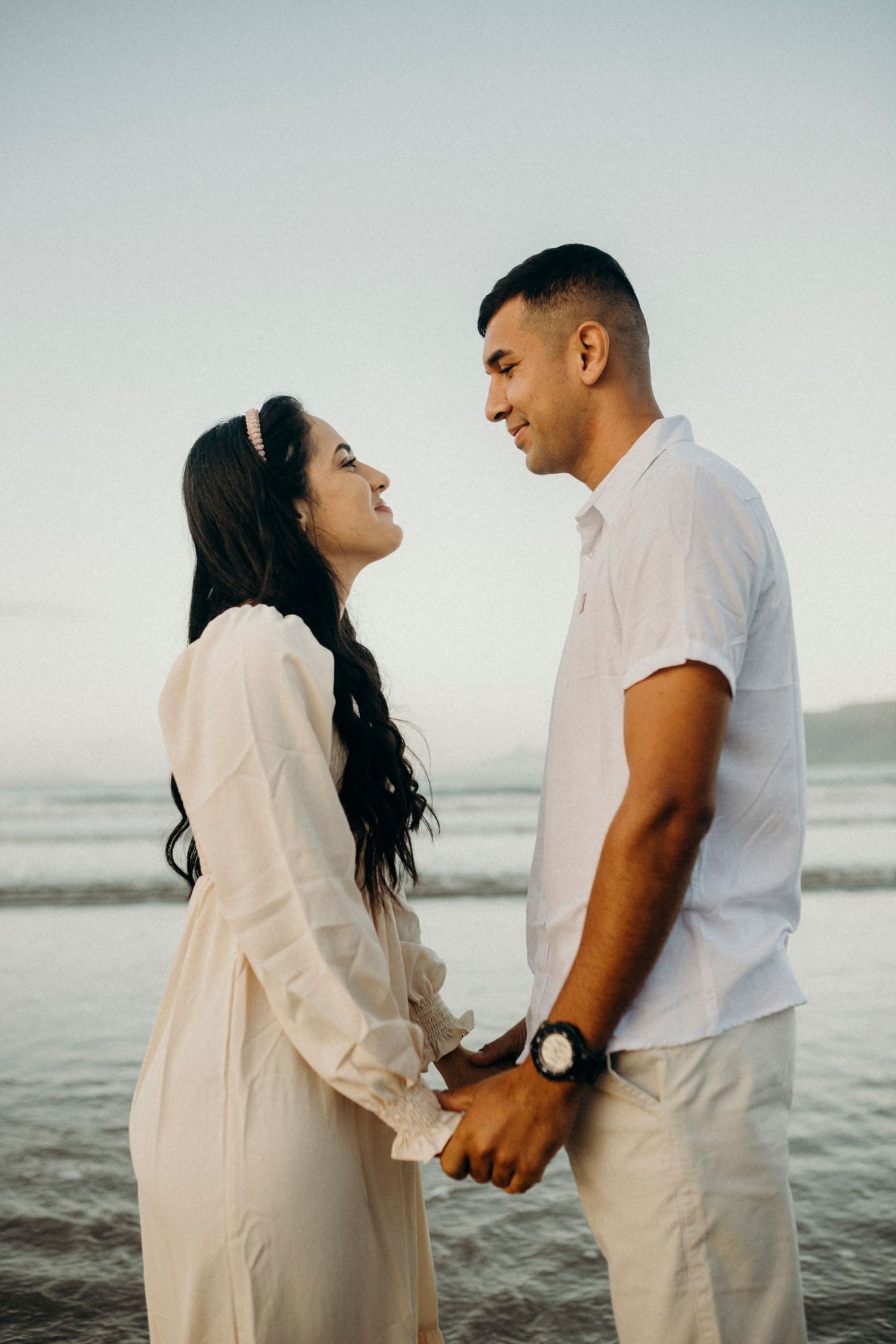 A romantic couple in white clothing shares tender moments during a beach photoshoot.