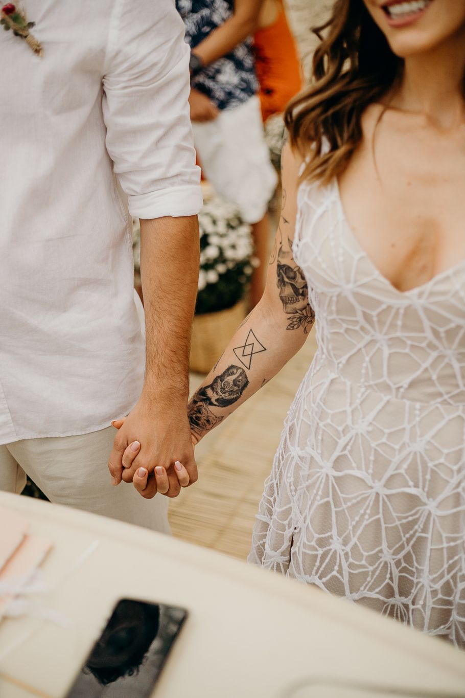 Wedding couple's hands shown with decorative tattoos and intricate lace dress details.