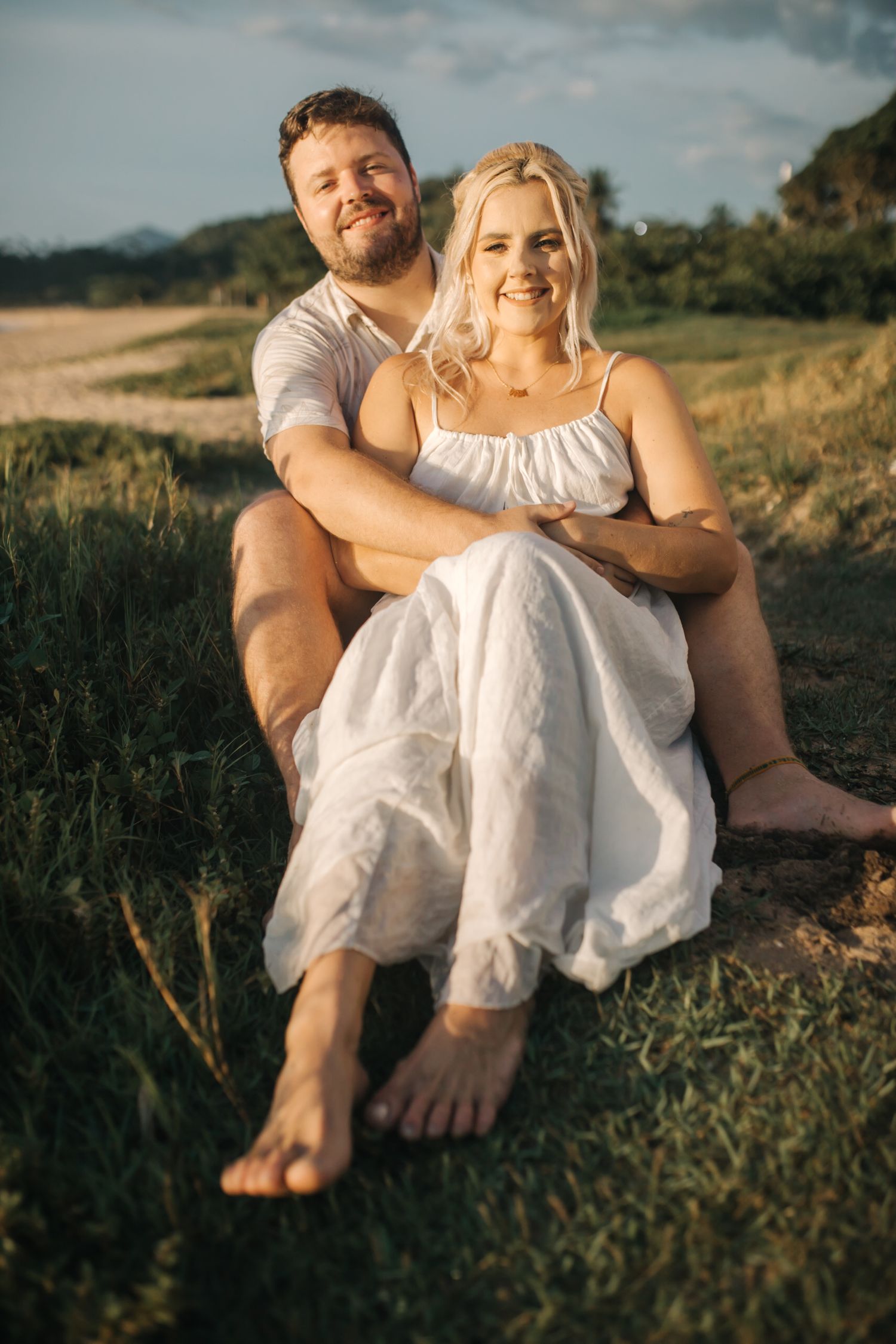 A casual outdoor portrait of a couple sitting together on grassy terrain at sunset.
