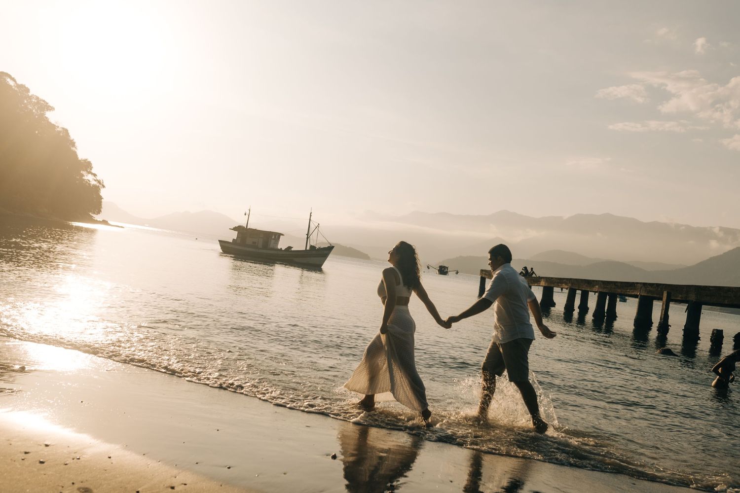 Silhouettes walking hand in hand along shoreline during sunset with boats in distance.